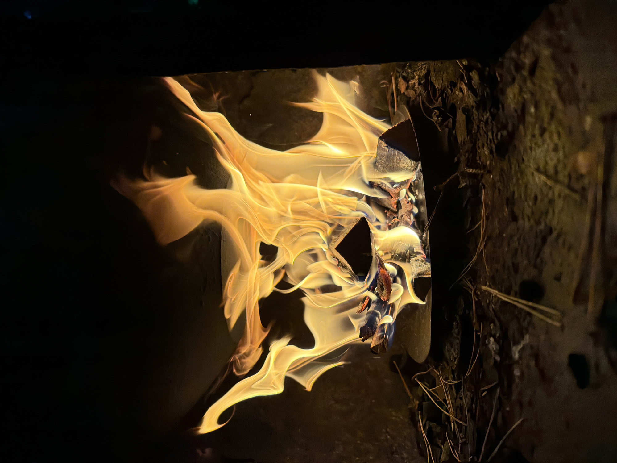 The image shows a close-up view of a fire burning in a fireplace or wood stove. The flames are predominantly yellow and orange, with some white at the hottest points. Several pieces of wood are visible, partially consumed by the fire. The background is dark, suggesting the surrounding environment is dimly lit or black. Scattered on the hearth are dry pine needles and other debris. The scene appears to be indoors, possibly in a residential setting. There is no indication of the location being in the city of Дмитровское, Russia.