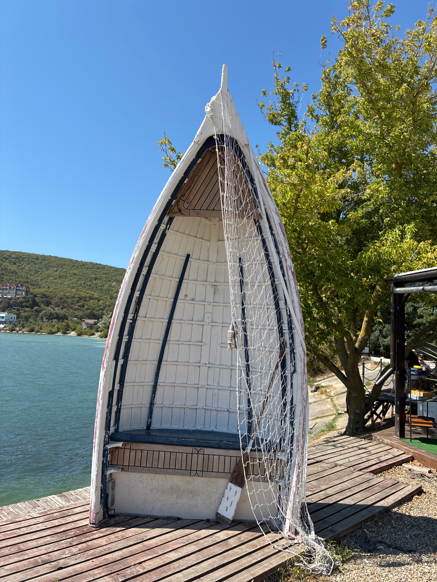 An inverted, white wooden boat with a wooden interior and a fishing net draped over its entrance sits on a wooden pier near the water in Abrau-Dyurso, Russia. The boat is positioned on a boardwalk constructed of weathered wooden planks, with a body of calm, turquoise water to its left. In the background, a lush green hillside dotted with buildings rises behind the water. A large tree with green foliage stands to the right of the boat, casting shadows on the pier. A small outdoor seating area with a dark roof structure is visible behind the tree. The sky is clear and blue.