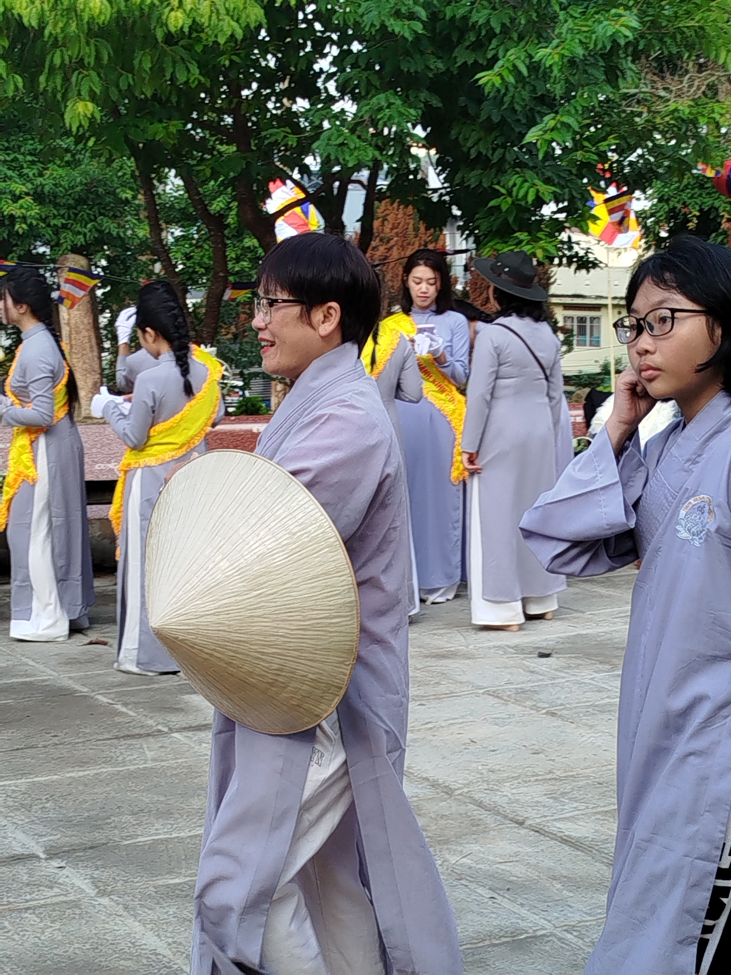In Da Lat, Vietnam, a group of people, predominantly women, are gathered in an outdoor setting paved with rectangular stone tiles. The women are dressed in light purple, traditional Vietnamese Áo Dài garments with white pants and some wear yellow sashes. One woman holds a Vietnamese conical hat. Some wear glasses. Multicolored Buddhist flags are strung overhead. A building is visible in the background.