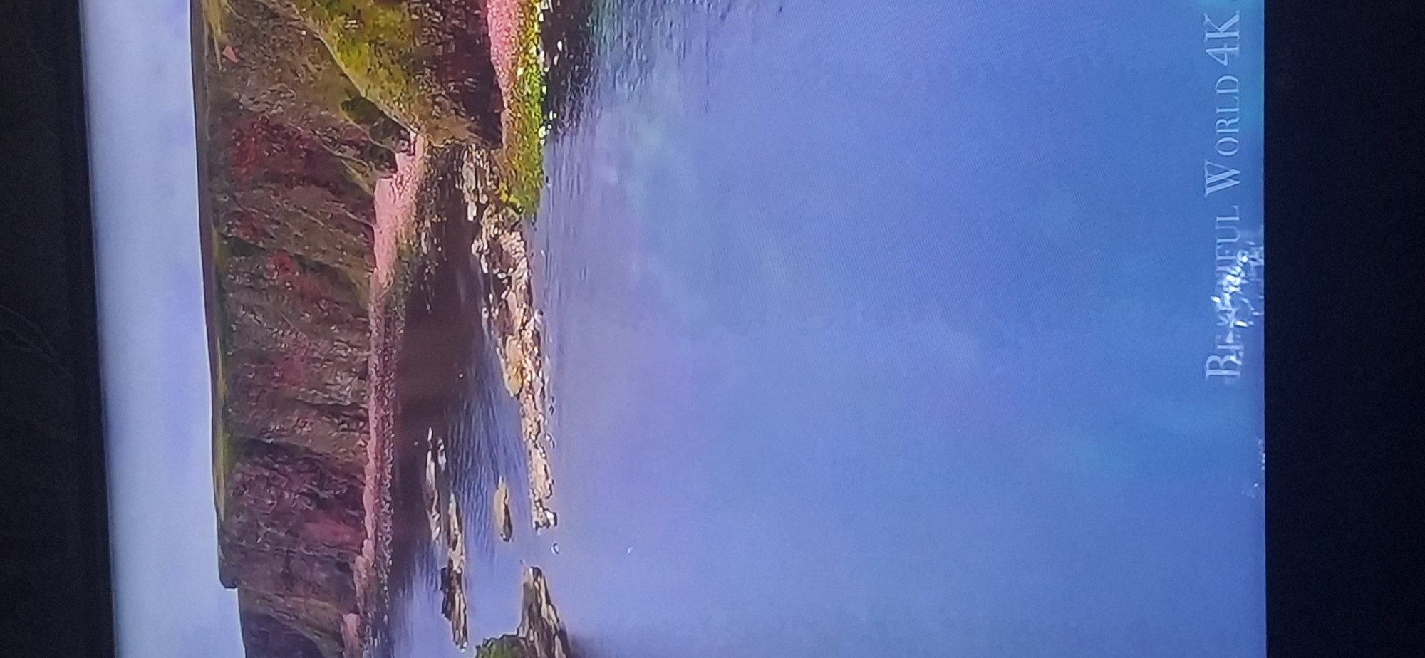 The image shows a coastal cliffside with the ocean in the foreground. The cliffs are a reddish-brown color, with patches of green grass on top. The water is a deep blue and there are several rocky outcrops visible in the water near the shore. The sky is a light blue. At the bottom of the image, the text "BEAUTIFUL WORLD 4K" is visible. The location is not Bila Tserkva, Ukraine.
