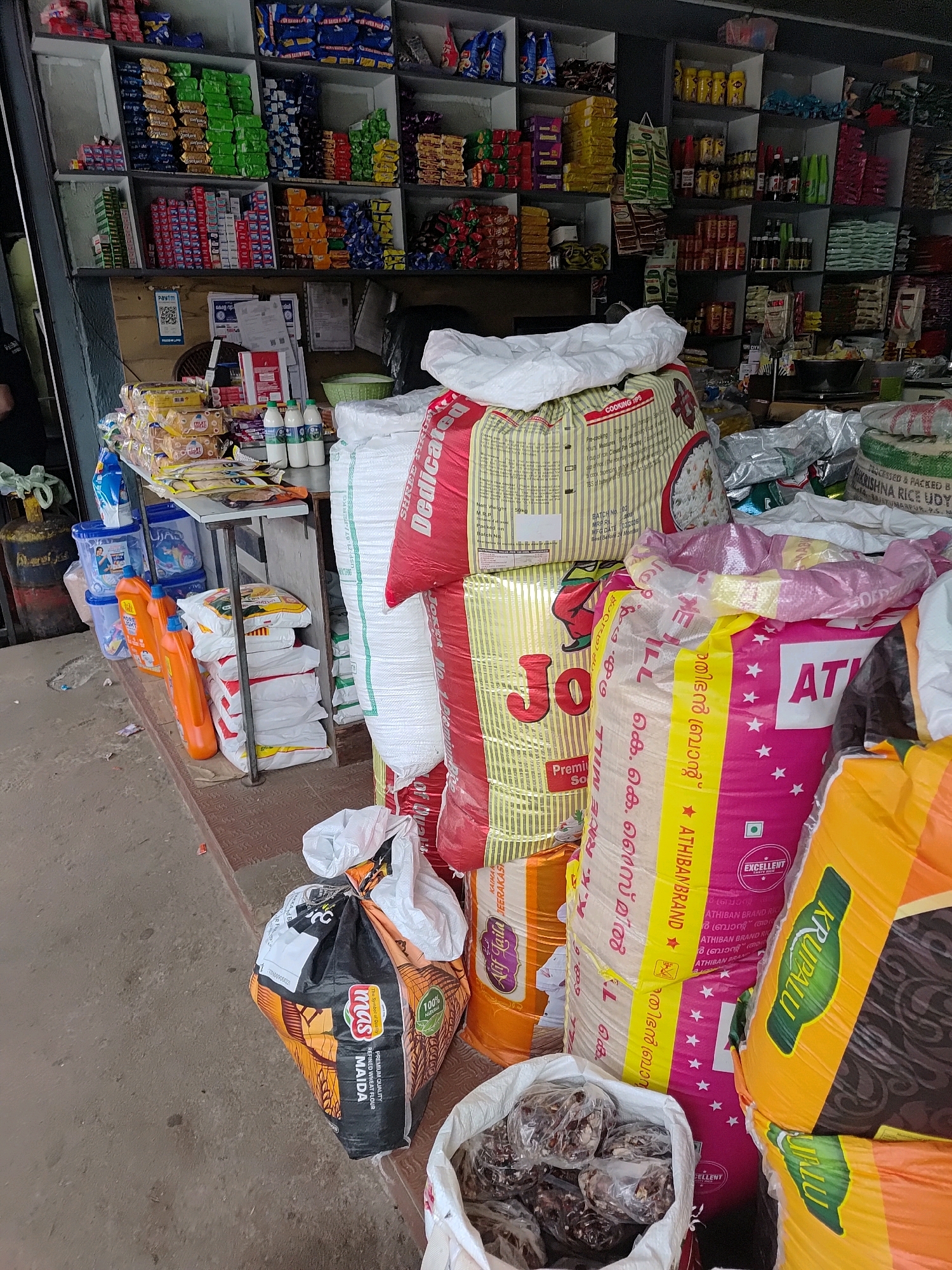 The image depicts a street-level view of a small grocery store or kirana shop, likely located in Mannarkkad, India. The foreground is dominated by a large assortment of stacked bags of rice and flour, with some smaller bags and containers of other food items also visible. There are large sacks of rice with colorful branding, including "JO" and "ATHIBAN BRAND". A large bag of "MAIDA" (refined wheat flour) is also prominent. To the left, there are several bottles of cleaning liquid and plastic containers. Behind these items, a shelf is laden with various packaged goods, including biscuits, snacks, and small boxes, arranged in neat rows. The background reveals more shelves stocked with a wider variety of products, suggesting a well-stocked retail space. The overall impression is one of a typical Indian grocery store with a dense display of goods. There are no people clearly visible in the frame. The environment appears to be an outdoor or semi-outdoor setting, with a concrete floor and a partially visible metal structure supporting the shelves.