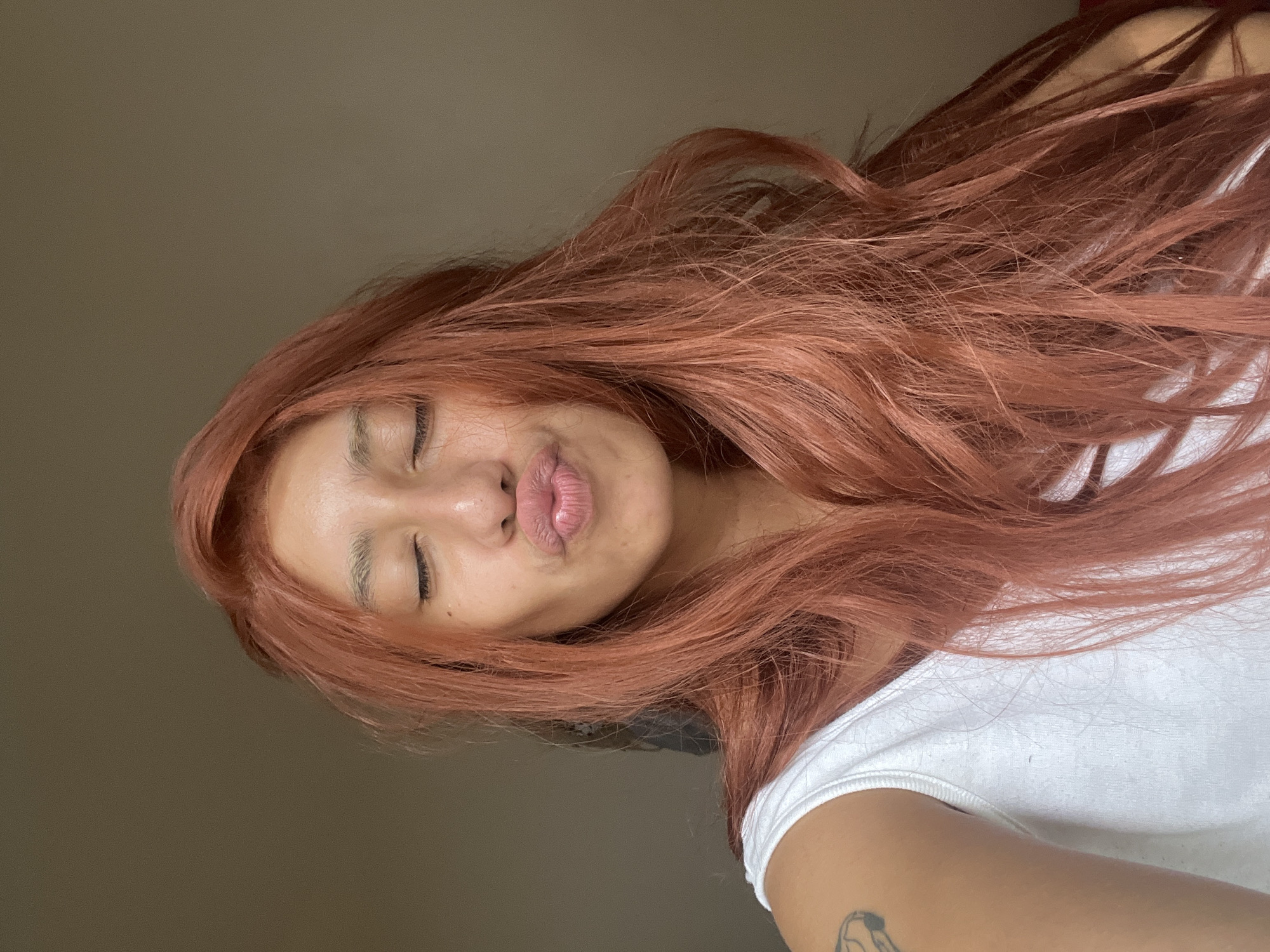 A close-up, slightly angled portrait captures a person, likely female, posing indoors. The subject has long, flowing reddish-brown hair that covers much of the frame, cascading over her shoulders. Her eyes are closed, and her lips are pursed in a playful "kissing face" or "duck face" expression. Her skin has warm undertones, and a slight sheen is visible on her forehead. She is wearing a simple white t-shirt, and part of her right arm is visible, revealing a black outline tattoo that appears to be an abstract or stylized design. The background is a plain, light brown or beige wall, suggesting an indoor setting without specific environmental cues. The overall mood conveyed is lighthearted and casual. There is no visible text in the image, and the time of day or specific location cues beyond being indoors are not discernible. The location context provided is Buenos Aires, Argentina.