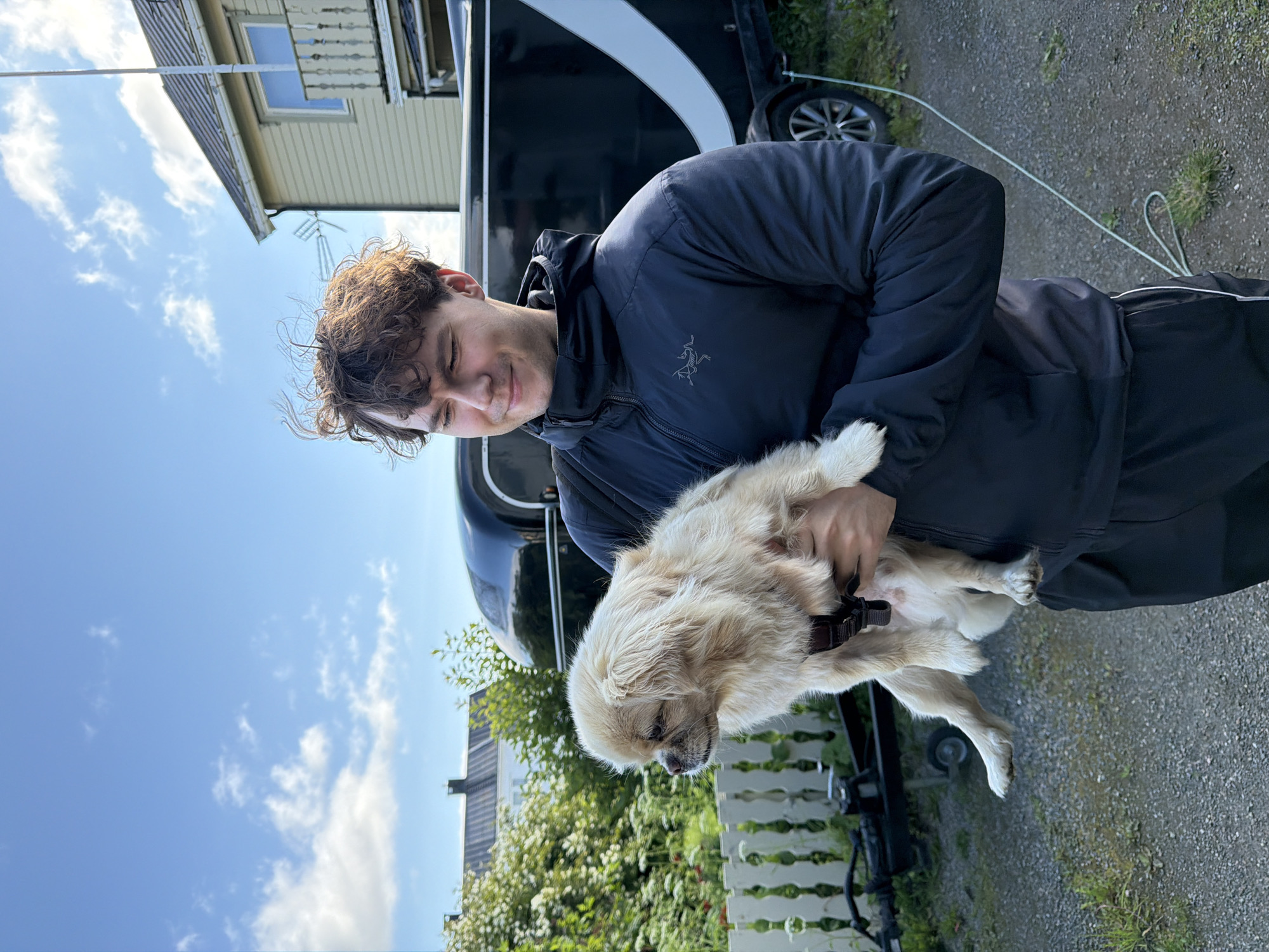 A young man with curly brown hair and a slight smile holds a small, fluffy, light-colored dog in his arms. He is wearing a dark blue hooded jacket with a zipper and black sweatpants with white trim. The dog is looking to the left. Behind them, there is a black trailer with a white stripe, a white picket fence, lush green bushes with white flowers, and a two-story building with a light green facade and white trim. The sky is blue with scattered white clouds. The scene appears to be outdoors in a residential area, likely in Harstad, Norway, given the architectural style of the building and the vegetation.