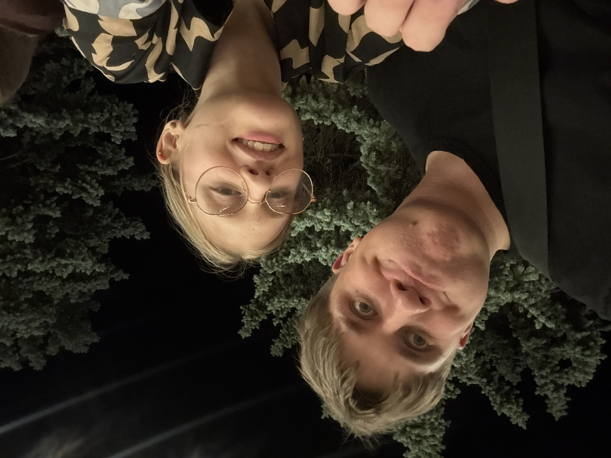 A medium shot captures a young couple taking a selfie at night, with lush green pine trees and a dark sky forming the background. The man on the left, with short, light brown hair, makes a comical face, puffing out his cheeks. He wears a black t-shirt. To his right, the woman, with blonde hair and round, gold-rimmed glasses, smiles at the camera. She is dressed in a black and beige patterned shirt. Their proximity suggests an intimate or friendly relationship. The lighting appears to be artificial, casting a soft glow on their faces and highlighting the textures of the pine needles. The scene is outdoors, likely in a park or a residential area with substantial greenery. The composition is informal, typical of a spontaneous selfie. The lack of specific landmarks makes pinpointing the exact location within Tiraspol, Moldova, challenging based solely on the visual information provided.