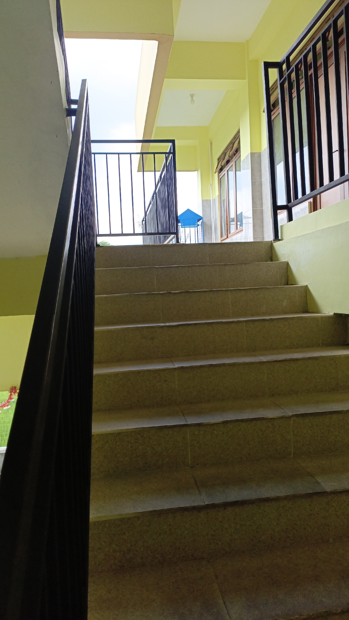A low-angle shot captures a set of tiled outdoor stairs leading upwards, partially framed by a dark railing on the left. The stairs are illuminated by natural light and appear to be made of light brown tiles. To the right, a yellow-painted wall runs parallel to the stairs, with dark metal railings and windows visible further up. In the background, a bright blue triangular structure stands against a light sky. The scene suggests a public building or pathway in Trihanggo, Indonesia.