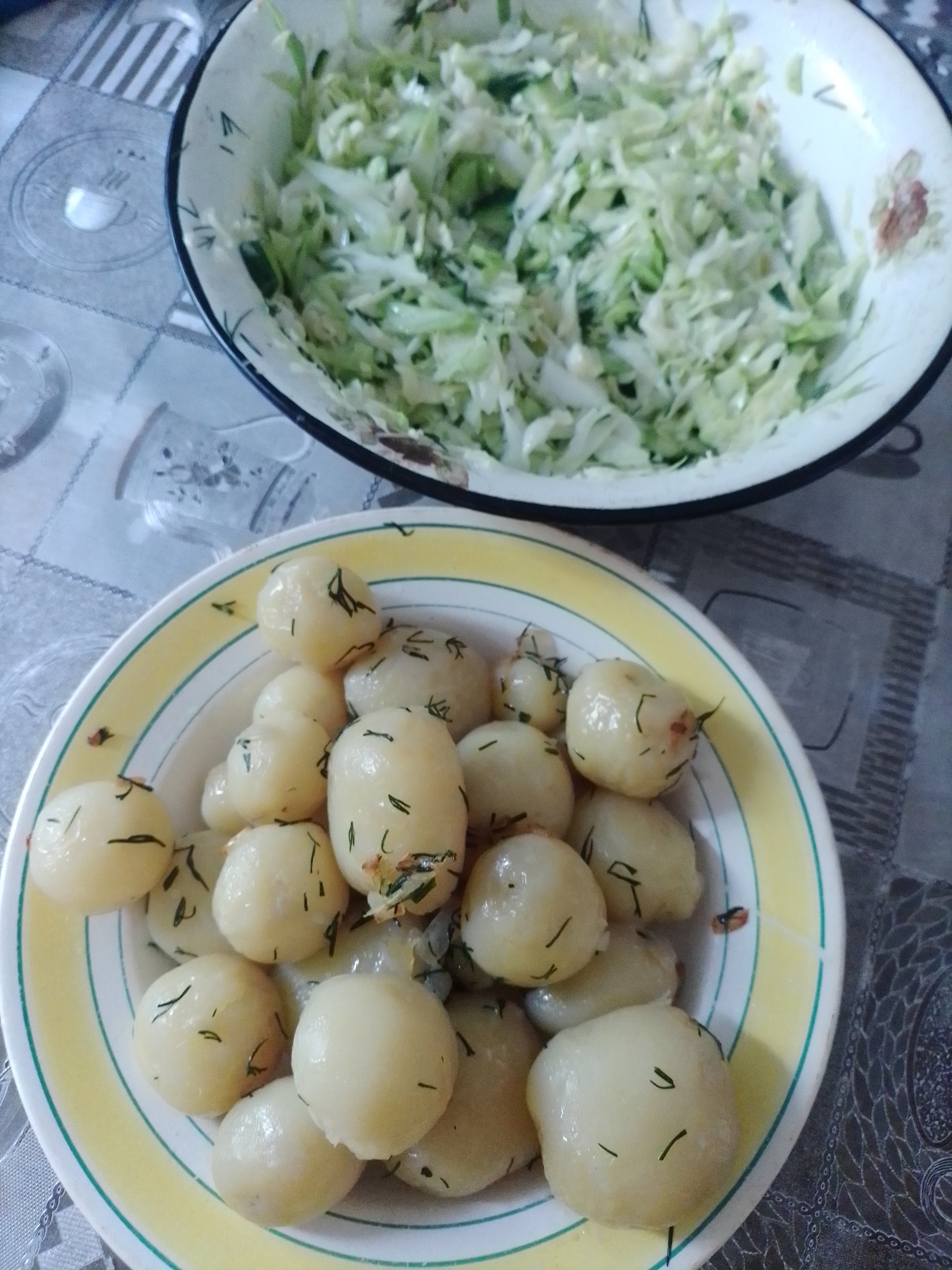 A close-up, eye-level shot shows two bowls of food on a patterned tablecloth. In the foreground, a white bowl with yellow and green stripes contains a pile of small, boiled potatoes. The potatoes are oval-shaped and appear to be seasoned with chopped green herbs, likely dill. Some potatoes have a glossy sheen, indicating they might be buttered or dressed. In the background, a larger, white bowl with a floral pattern holds a generous portion of shredded cabbage salad, mixed with what appears to be dill or other herbs. The tablecloth has a grey and white pattern resembling lace or a damask print, with some circular motifs visible. The scene is indoors, likely a dining setting in Ustyvytsia, Ukraine.