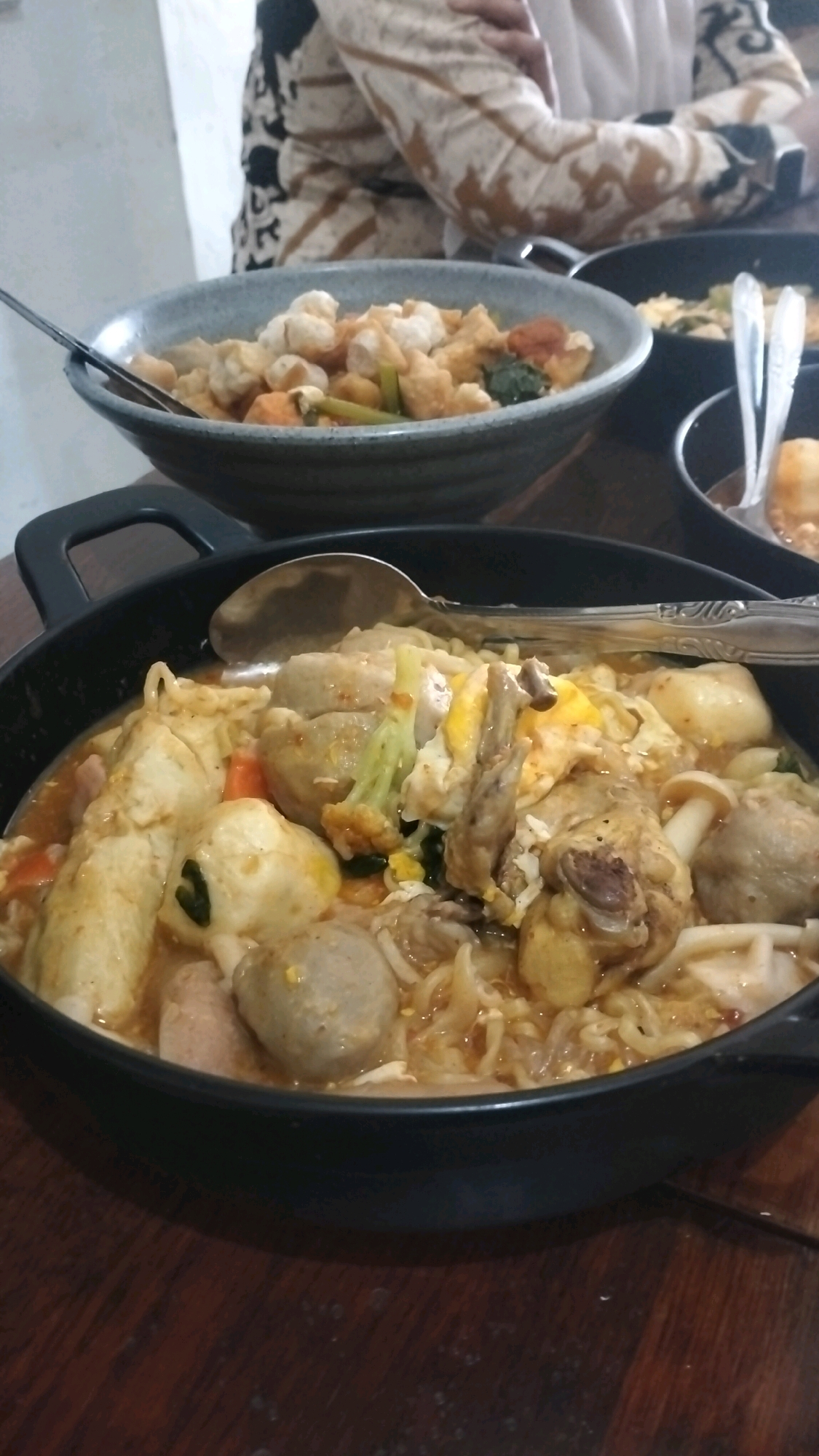 A close-up, overhead shot shows a black bowl filled with a hearty noodle soup dish. The soup contains various ingredients including meatballs, chicken pieces, broccoli florets, and possibly egg. A spoon rests on the edge of the bowl, slightly submerged in the broth, suggesting the meal is being eaten. In the background, out of focus, a person wearing a patterned garment is visible, seated at a table. The table surface is made of dark wood. The overall scene suggests a casual dining experience, likely in a restaurant in Lhokseumawe, Indonesia, given the context provided.