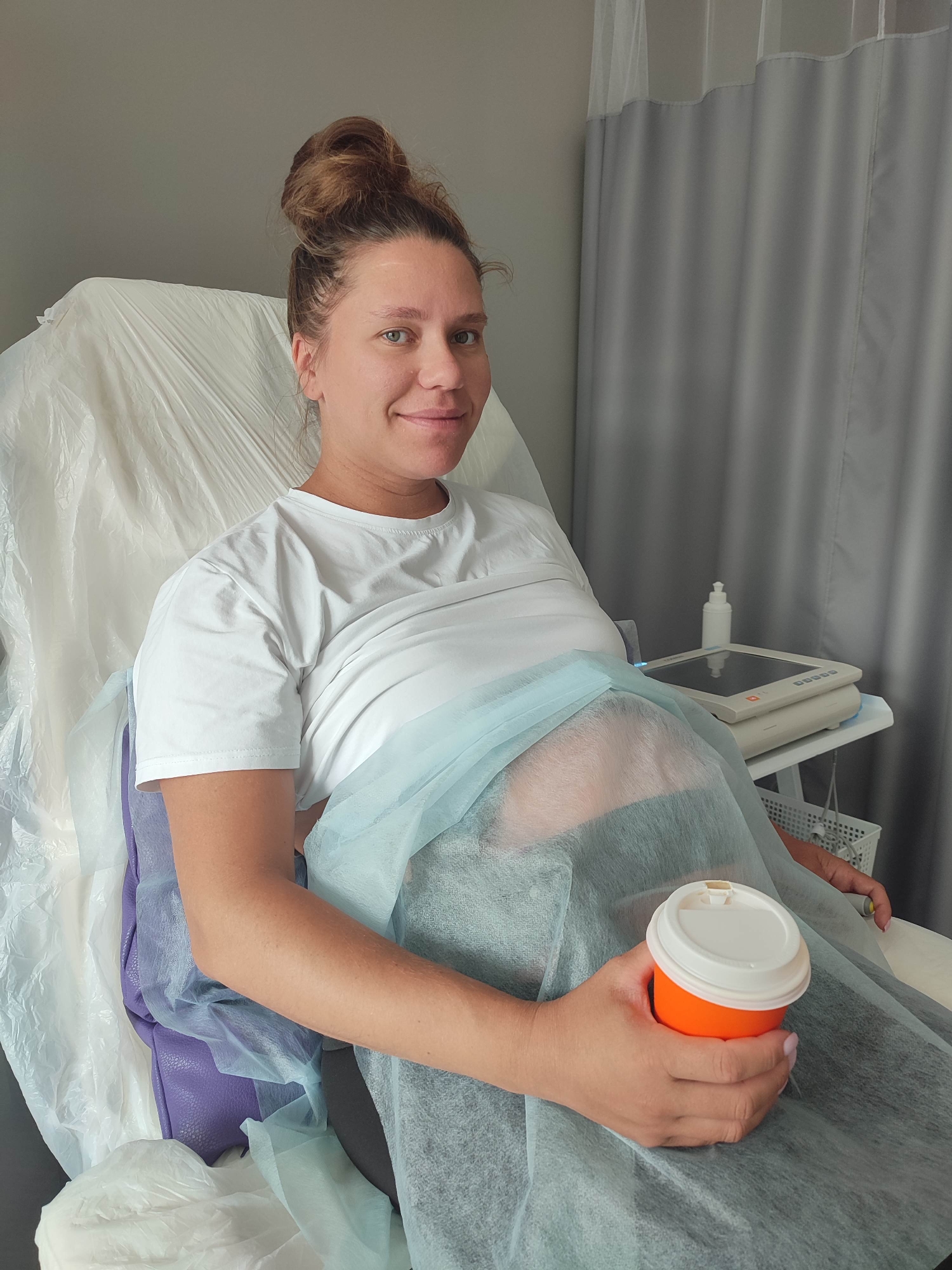 A pregnant woman with her hair in a bun is sitting in a medical examination chair, wearing a white t-shirt and covered by a blue medical drape. She is holding an orange and white disposable cup in her right hand. A medical ultrasound machine is visible on a stand to her right. The setting appears to be a medical clinic or hospital room, with gray curtains in the background. There is no identifiable information indicating this location is in Izhevsk, Russia.