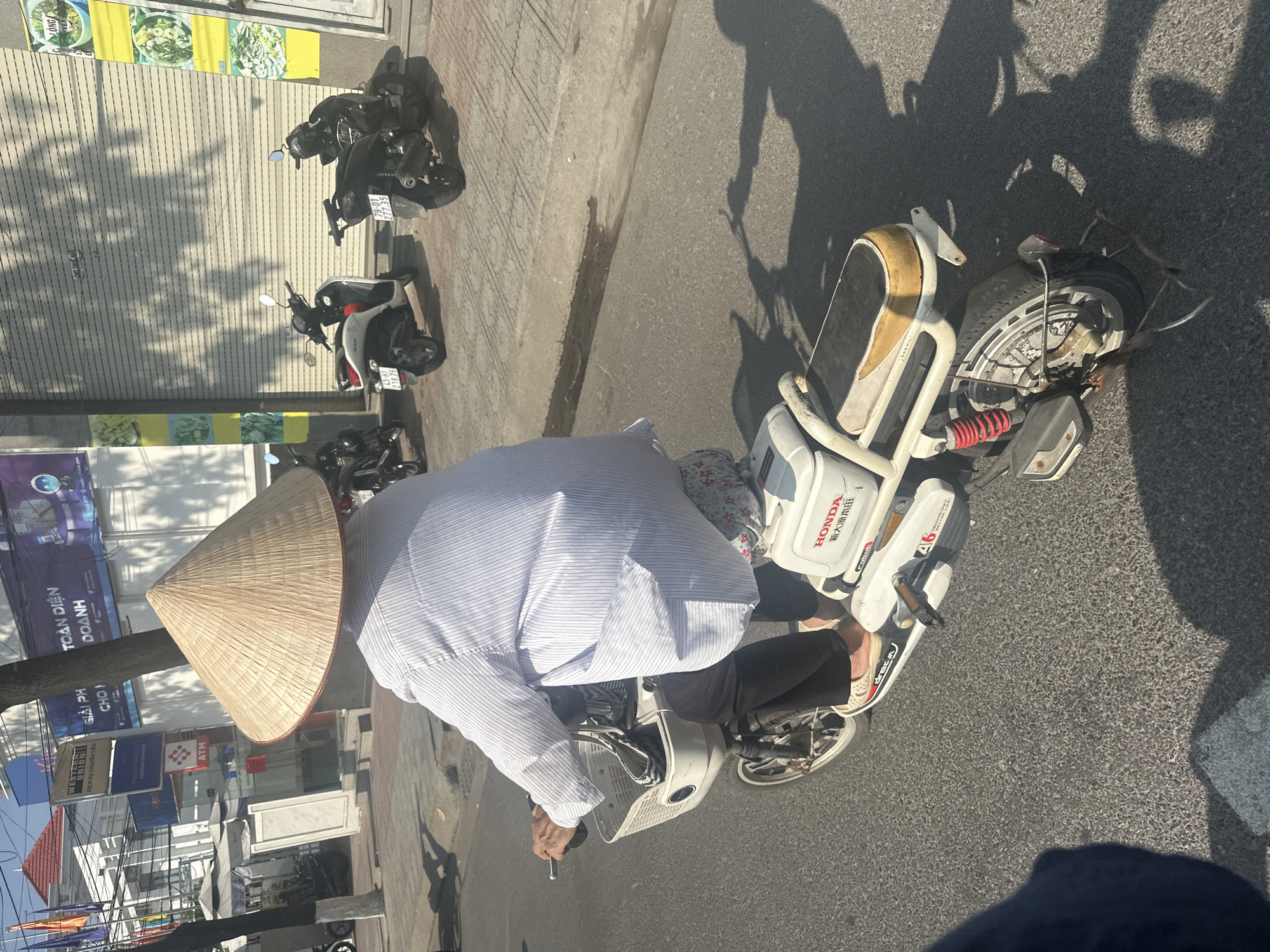 A high-angle, eye-level medium shot shows a man wearing a traditional Vietnamese conical hat, a light blue and white striped long-sleeved shirt, and dark pants, riding a white and beige Honda electric scooter on a street in Bac Nha Trang Ward, Vietnam. The man is seen from the rear, his posture suggesting he is driving. His hands are visible on the scooter's handlebars.
Several parked motorbikes of various colors and models are visible to the left, along the edge of the sidewalk. In the background, shopfronts with signage in Vietnamese are visible. The street is paved with grey asphalt. Shadows of the man and other objects are cast on the road, indicating strong sunlight.