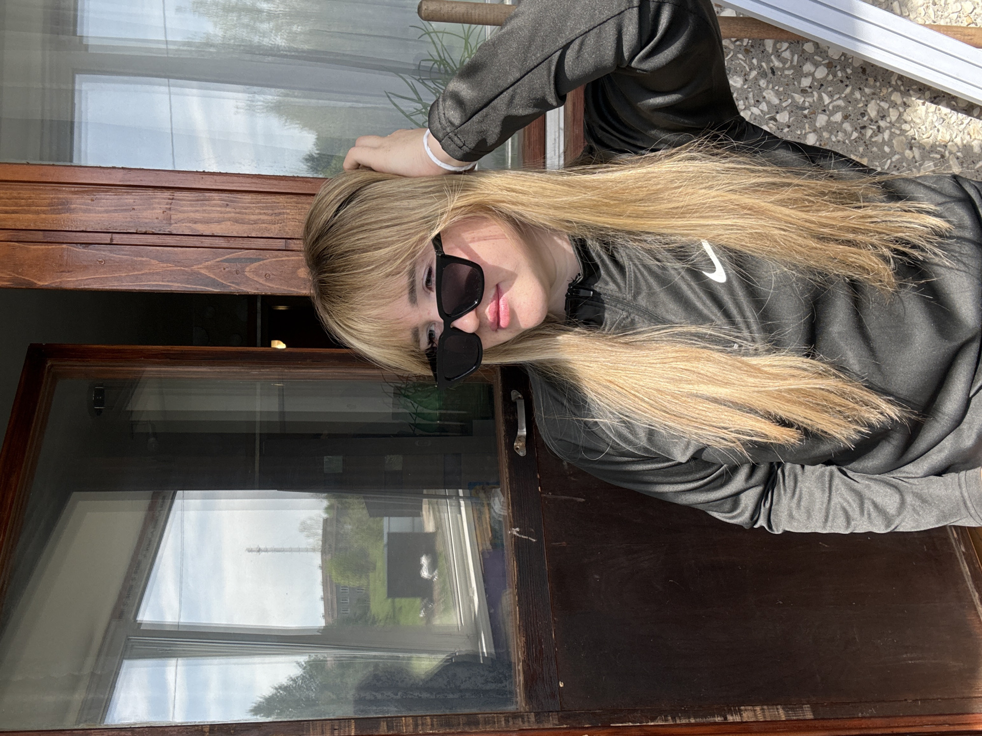 A woman with long, blond hair and sunglasses is leaning on a dark-stained wooden window frame. She is wearing a dark-gray, long-sleeved shirt with a white Nike swoosh on the chest and a white bracelet on her wrist. The reflection in the window shows a rural landscape with a structure resembling a tower. Given the location of Марковский, Russia, based on previous context, the landscape appears to be consistent with the region's rural areas. The surface she is leaning on appears to be a wooden window frame.