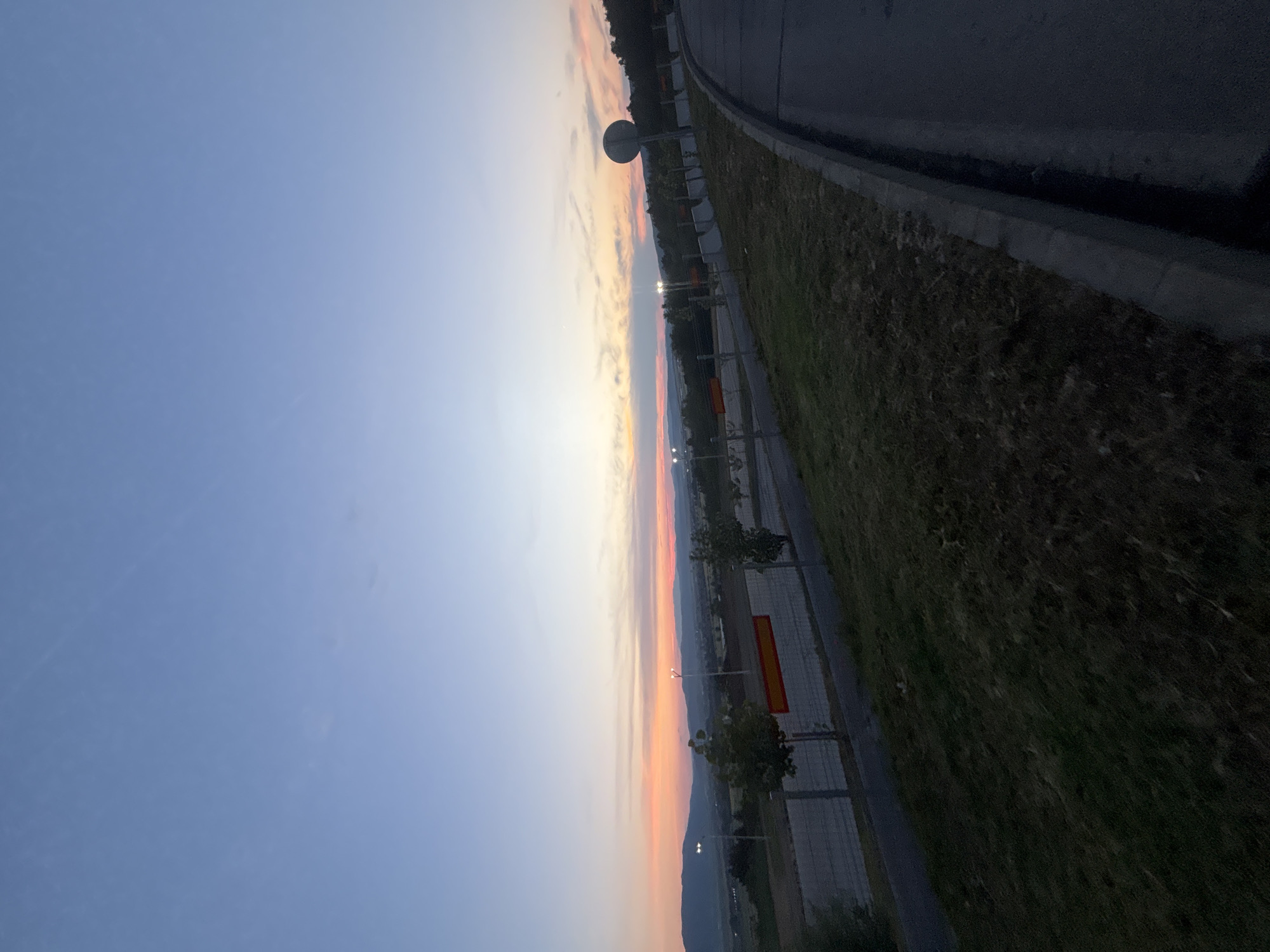 The image depicts a twilight scene in Ihtiman, Bulgaria, likely at dusk. A road curves into the foreground on the right, with a grassy embankment on the left. A concrete curb separates the road from the grass. In the midground, a fence runs parallel to a paved path. Beyond the fence, there is a wide expanse of land with trees and what appears to be a sports field or stadium, indicated by a long red and yellow banner on a fence. In the distance, rolling hills form the horizon under a gradient sky transitioning from a soft orange and pink near the horizon to a pale blue overhead. Two streetlights are visible, one to the left and one more centrally located, casting a soft glow. A circular traffic sign is also present on the right side of the image, positioned on a pole. The overall atmosphere is serene and peaceful, characteristic of the transition from day to night.