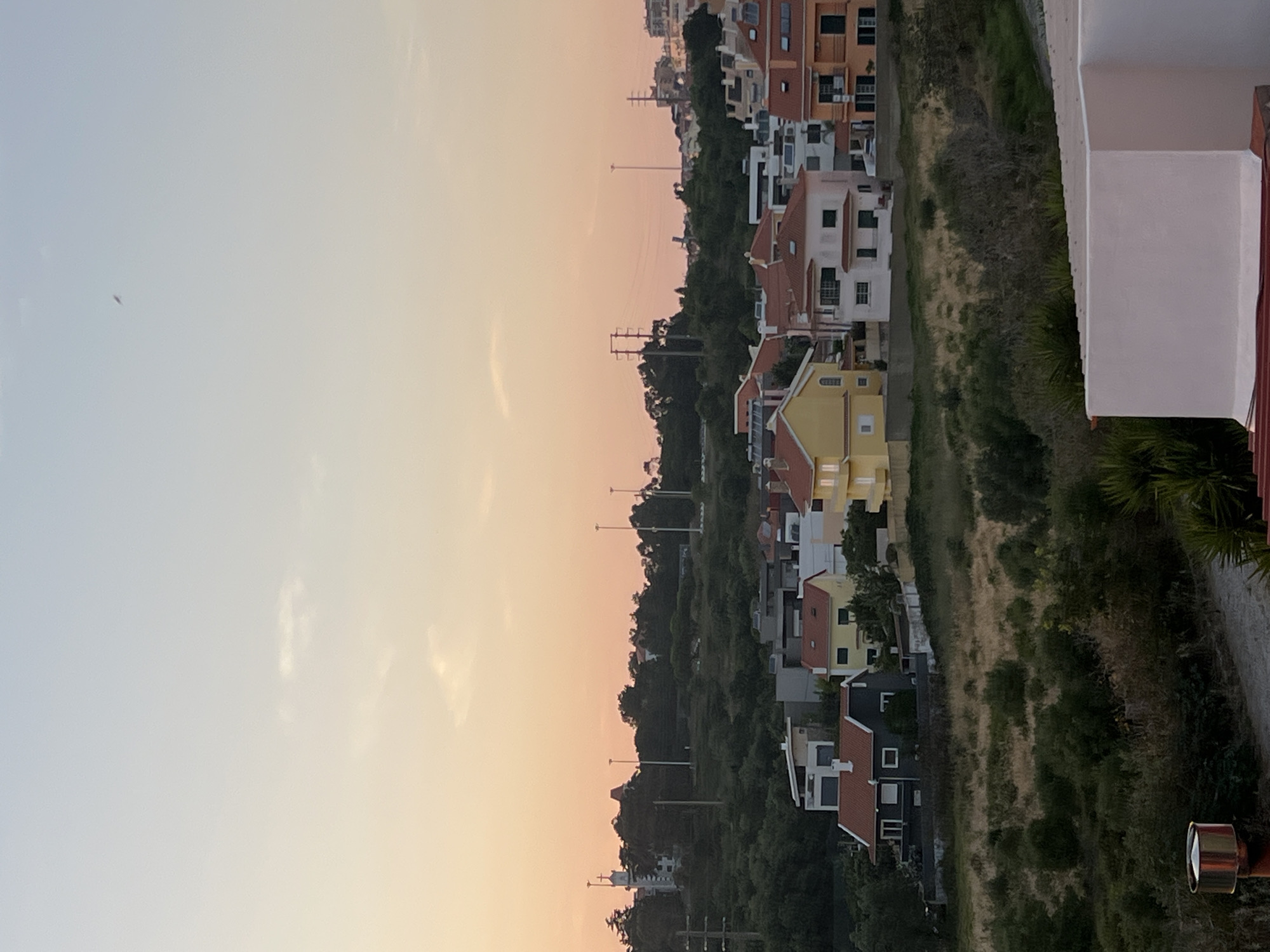 This is a full, eye-level, outdoor, vertical shot of a residential area on a hill in Cascais, Portugal, at dusk. The sky is a gradient of pale blue and soft orange, with a few wispy clouds. In the distance, a small dark object is visible in the sky.
The middle ground features a cluster of houses with red-tiled roofs. Some houses are painted white, others yellow, and one is dark gray. They are situated on a grassy, overgrown slope dotted with bushes and trees. Further up the hill, more houses and some taller buildings are visible, interspersed with trees and utility poles. A white building with a cross on top, possibly a church, is also visible on the higher part of the hill.
The foreground shows a rooftop or balcony railing of a white structure, with a glimpse of a red-tiled roof below it. To the left, a metal object, possibly a chimney or ventilation unit, stands near a textured pathway. The overall scene depicts a peaceful, twilight view of a suburban Portuguese landscape.