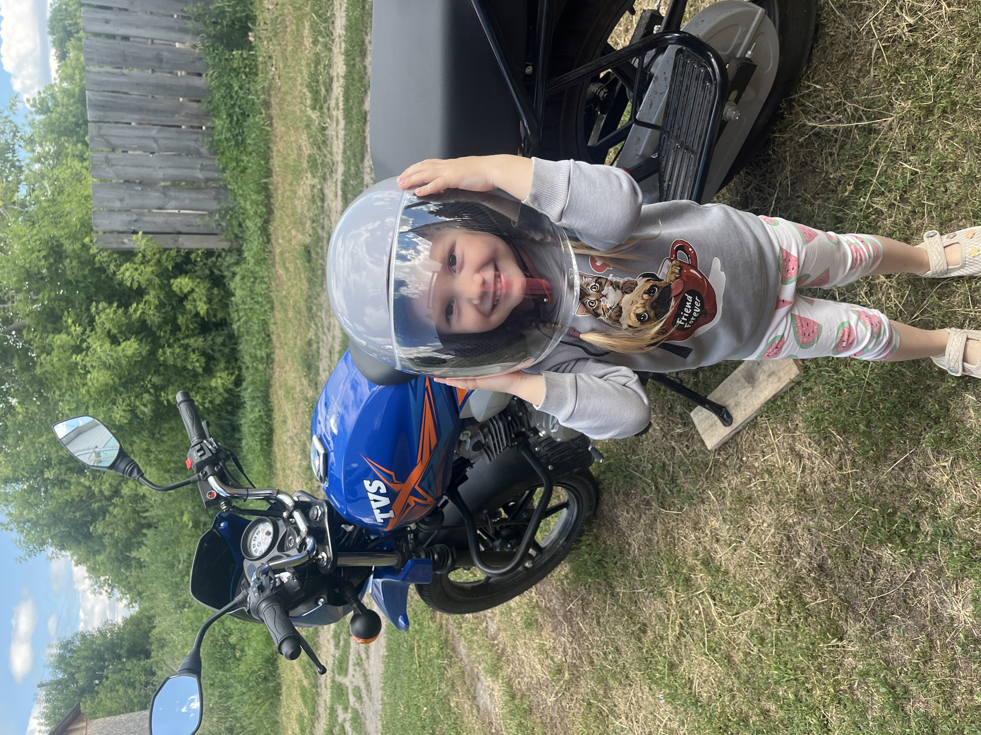 A young girl wearing a clear helmet and a gray sweatshirt with a dog and cat graphic is posing with a blue and orange motorcycle. She is smiling and holding the helmet on her head. The motorcycle has "TVS" written on its side. She is wearing white pants with a watermelon pattern and light-colored sandals. The scene is outdoors on a grassy area, with a wooden fence and trees visible in the background. The lighting suggests it is daytime, likely sunny. The setting appears to be rural, consistent with the provided location context of Сухринское, Russia. The girl's expression conveys joy and excitement.
