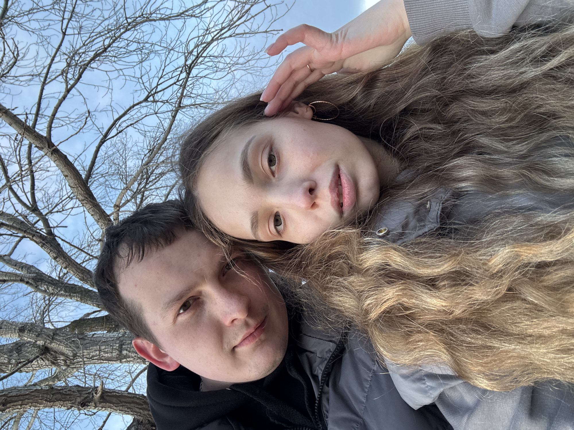 A selfie-style photograph shows a man and a woman close together outdoors. The man, with short dark hair, is wearing a black hooded jacket. The woman has long, wavy, light brown hair and is wearing a gray jacket. Her hand is raised to her forehead, and she is wearing hoop earrings and a ring. The background consists of bare tree branches against a light blue sky. The location is Chaikovsky, Russia.