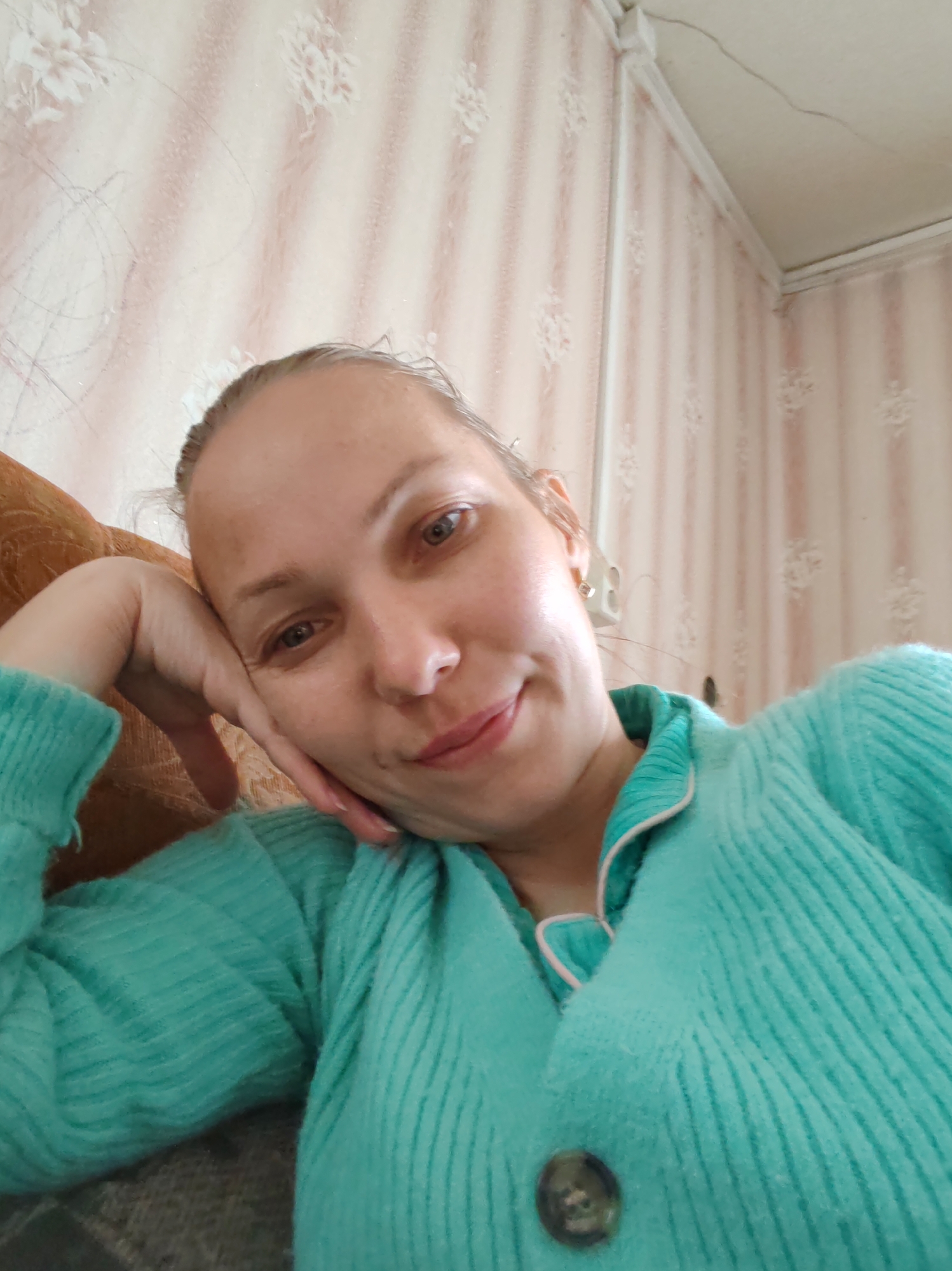 This is a close-up, slightly upward-angled selfie of a woman indoors, likely in Чухлэм, Russia. She appears to be in her 30s or 40s, with light-colored hair pulled back from her face, revealing a high forehead. Her eyes are light-colored, possibly hazel or light brown, and she has a gentle, closed-mouth smile. Her right hand is propping up her head, resting against her cheek and chin, with her arm visible.
She is wearing a soft-looking, ribbed cardigan or top in a vibrant turquoise or teal color. The garment has a light pink or white piping along the collar and front opening, with a visible dark, round button near the bottom center of the frame.
The background consists of a wall covered in wallpaper with a light beige or off-white base featuring faint vertical stripes and scattered floral or leaf patterns in a lighter tone. Some darker, abstract scribbled lines are also faintly visible on the wallpaper, possibly wear or marks. In the upper right corner, the corner where two walls meet the ceiling is visible. There's a white ceiling molding or trim along the wall-ceiling joint, and a crack or seam is visible on the ceiling. A white electrical outlet or light switch is mounted on the wall behind her right ear. Part of a brown, upholstered sofa or armchair is visible behind her left shoulder and arm.
The lighting appears natural and soft, suggesting daytime. The overall mood is relaxed and calm. No visible text is present in the image.