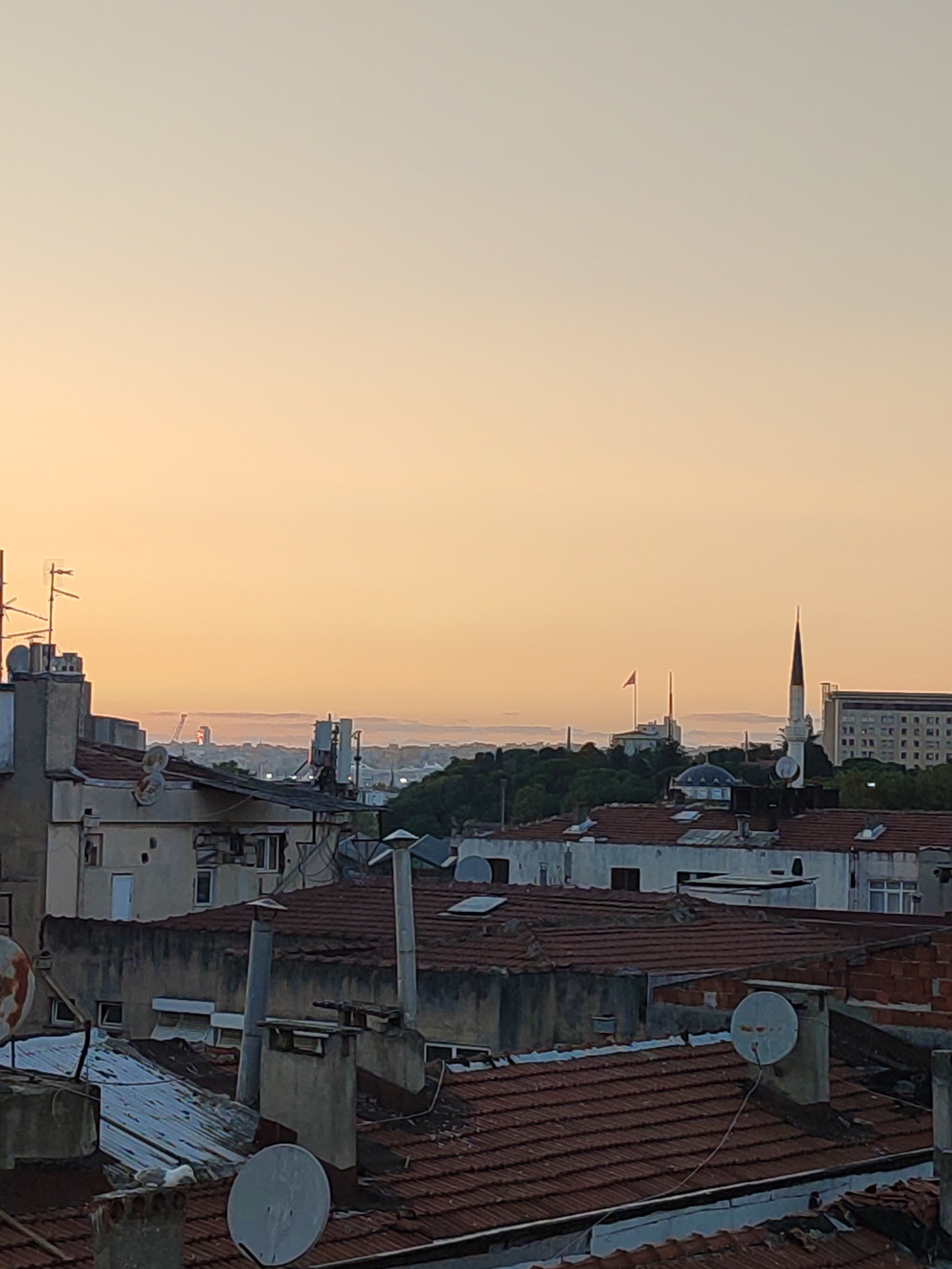 An outdoor, low-angle, medium shot captures a cityscape at dusk, likely in Kadıköy, Turkey, as indicated by the architectural style and the presence of a minaret. The sky is a soft gradient of pale orange and yellow, transitioning into a lighter hue towards the top of the frame, suggesting the setting sun.
In the foreground, numerous terracotta-tiled rooftops dominate the scene. These roofs are cluttered with various objects, including chimneys of different shapes and sizes, satellite dishes, and some metal sheeting. The buildings appear to be older, with some showing signs of wear and tear, such as peeling paint and weathered facades. Antennas are visible on some of the structures.
In the mid-ground, a collection of buildings forms a dense urban landscape. To the right, a white minaret with a slender spire stands prominently, a characteristic feature of Islamic architecture. Behind it, a larger, modern building with multiple windows is visible. Further left, two flagpoles with flags are erected on top of a building. A dense area of green trees forms a natural barrier between the foreground and the distant cityscape.
In the distance, a hazy skyline is punctuated by more buildings and what appears to be a construction crane. The overall impression is one of a lived-in, urban environment viewed from an elevated perspective as daylight fades. There are no visible people or animals in the image.