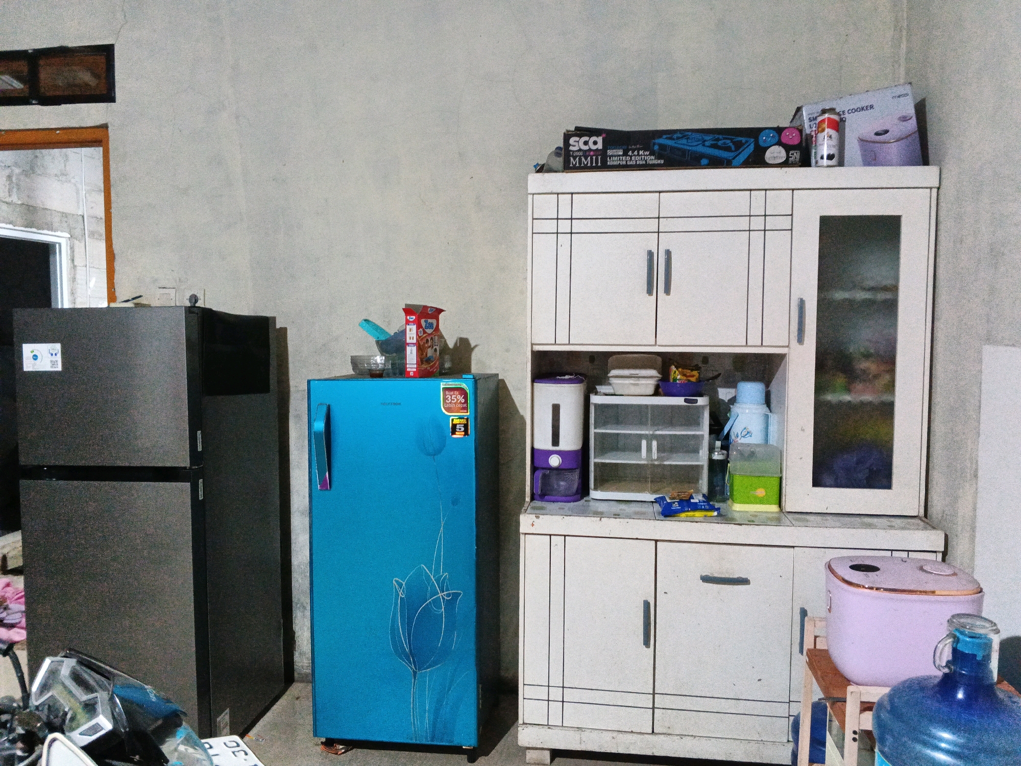 The image depicts the interior of a simple, somewhat cluttered room, likely a kitchen or utility area, in Sirau, Indonesia. On the left, a large, dark-colored refrigerator stands next to a bright blue refrigerator with a stylized tulip design. Above the blue refrigerator, a box of snacks is visible. To the right, a white cabinet unit with multiple shelves and drawers takes up a significant portion of the frame. On top of this cabinet, various items are arranged, including a blue gas stove box, a small white appliance, and a rice cooker. Below the cabinet, a small pink rice cooker and a large blue water jug are placed on the floor. The walls appear to be concrete, with some visible cracks. A small window is situated high on the wall to the left. The overall impression is of a functional, lived-in space.