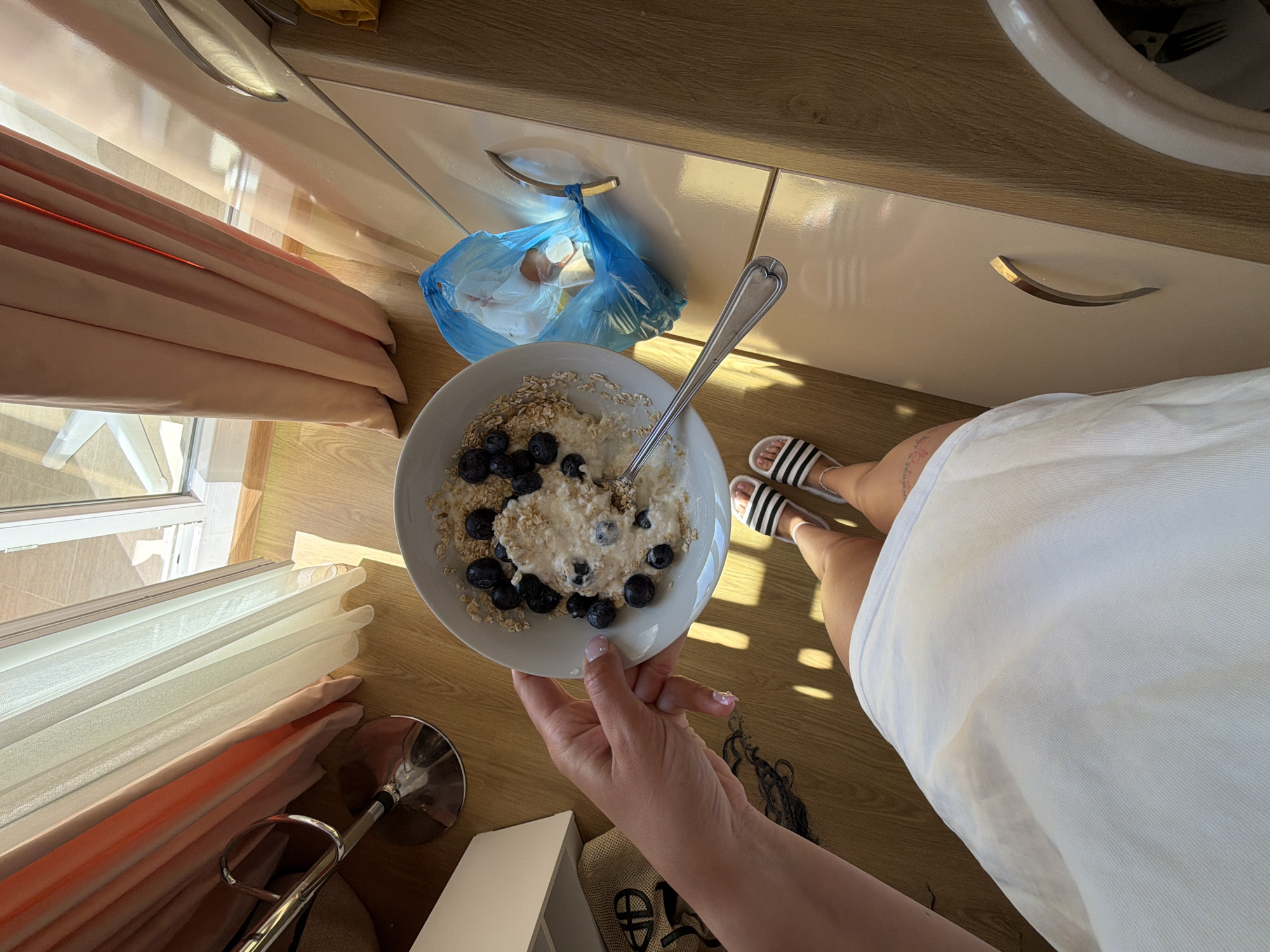 The image depicts a breakfast scene with a bowl of oatmeal topped with blueberries. A person's hand is holding the bowl. The person is wearing white slides with black stripes and a white dress. The setting appears to be an indoor space with wooden flooring and light-colored cabinets. Curtains and a window suggest a room with natural light. A blue trash bag containing eggshells is visible on the counter. The scene is set in Nesebar, Bulgaria.