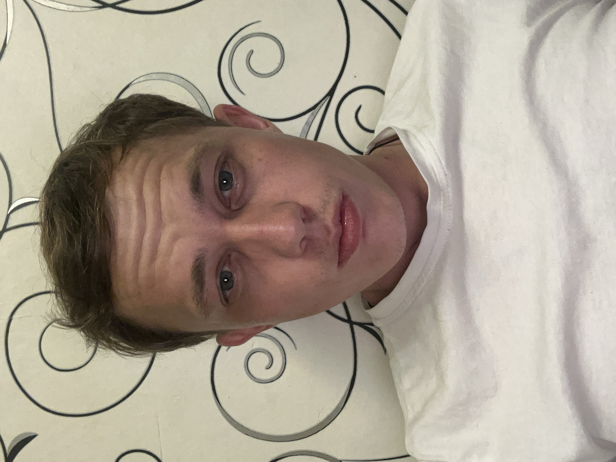 A medium shot shows a young Caucasian male with light brown hair and blue eyes looking directly at the camera. He is wearing a white t-shirt. The background is a wall with a light-colored wallpaper featuring a swirling black and gray pattern. The lighting is dim, and shadows are visible on his face. The image appears to be taken indoors. The city of Novosibirsk, Russia is not determinable from the image content.
