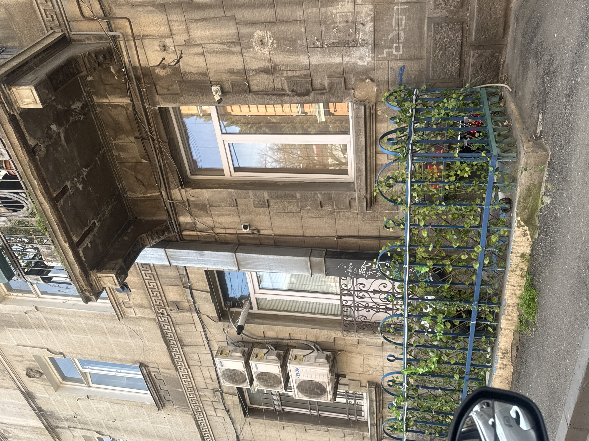 The image depicts a building facade in Tbilisi, Georgia, characterized by its stone construction. The building features windows of varying sizes and shapes, with some windowsills adorned with air conditioning units. An ornate blue metal fence, intertwined with artificial vines, runs along the ground floor adjacent to a street. The architectural details include a balcony with intricate metalwork, and decorative moldings are visible on the building's exterior. The street surface is adjacent to the building.