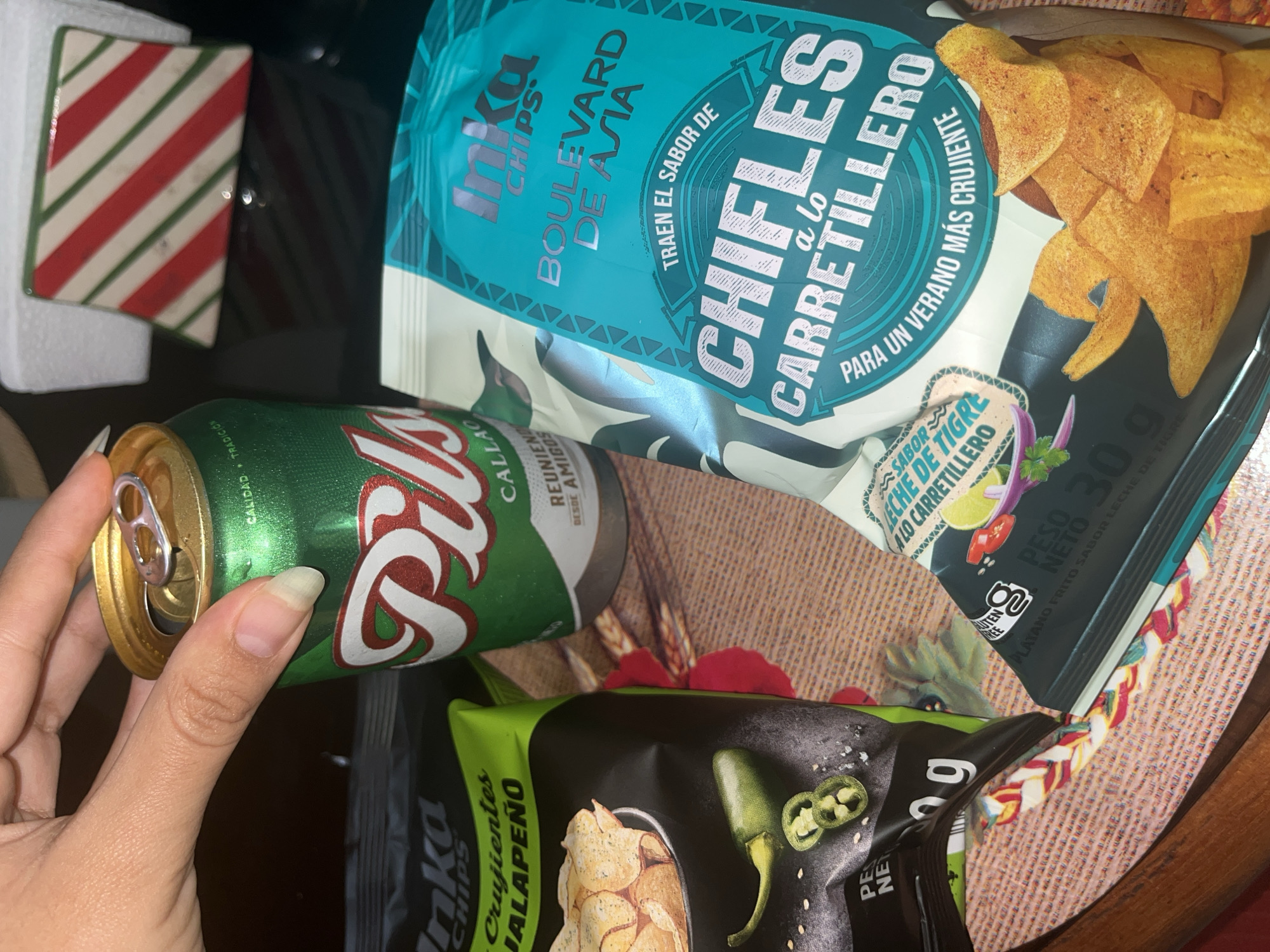 The image shows a hand holding a green and gold Pilsen Callao beer can. In the background, there are two bags of Inka Chips: one labeled "Boulevard de Asia Chifles a lo Carretillero," and the other labeled "Crujientes Jalapeno." The chips are on a patterned mat. There is also a square coaster with red and white stripes. The brand of beer, "Pilsen Callao" indicates a Peruvian location, specifically the city of Callao near San Luis, Peru.