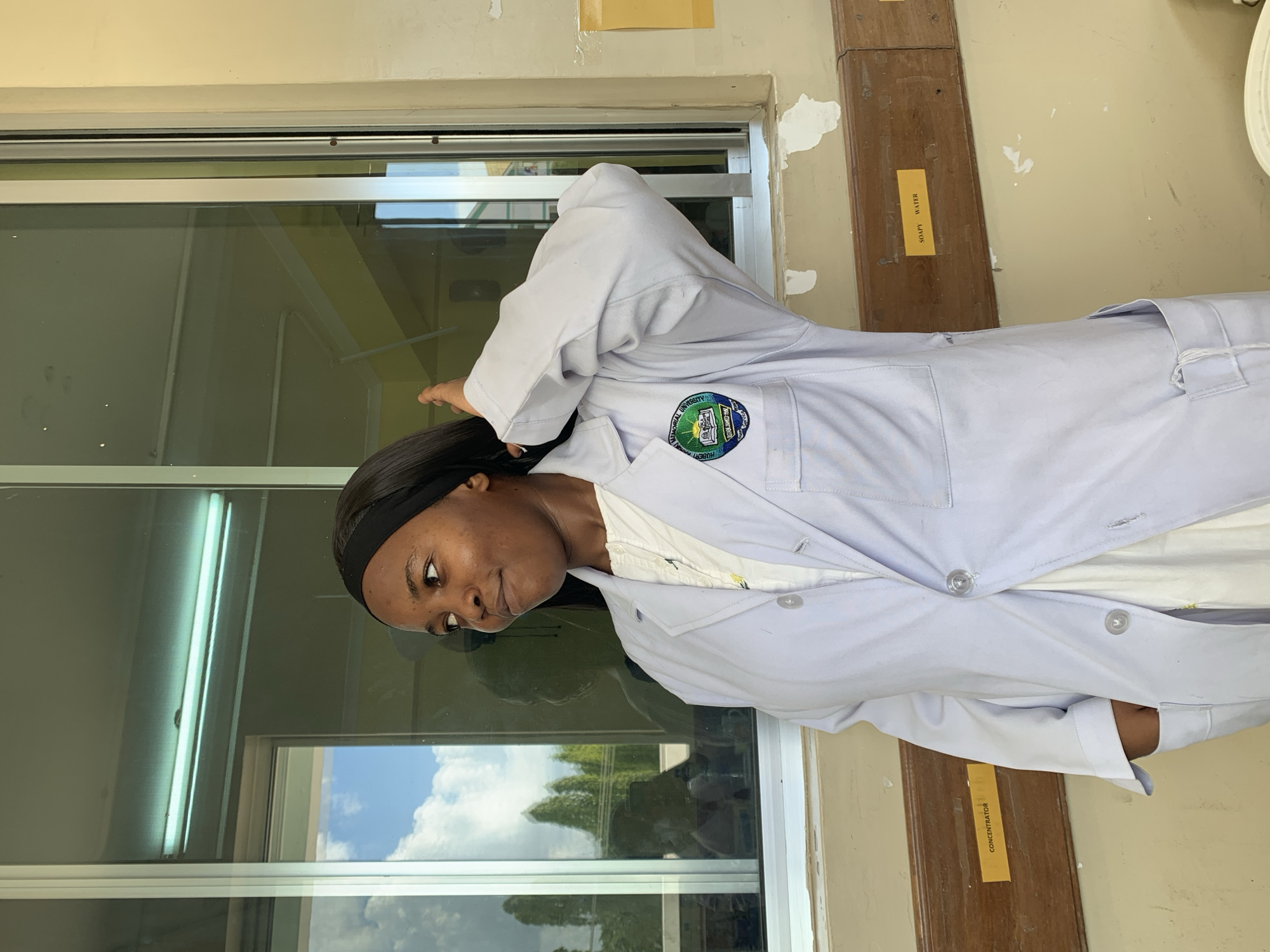 A young Black woman stands in what appears to be an interior space, likely a laboratory or educational facility. She is wearing a white lab coat with a circular emblem on the left breast area, possibly indicating a university or research institution, and a black headband. The lab coat is partially unbuttoned, revealing a collared shirt underneath.
She is positioned in front of a large window, which is reflecting an exterior scene of trees and sky, presumably an outdoor setting. The building's interior walls are painted in a pale yellow or off-white color. Adjacent to the window are wooden planks with labels attached, with the words "SOAPY WATER" and "CONCENTRATOR" visible.
While it is not possible to determine the exact location as Dar es-Salaam, Tanzania, solely from the image, if one assumes she is associated with the Hubert Kairuki Memorial University in Dar es-Salaam, the location could be related to this institution.