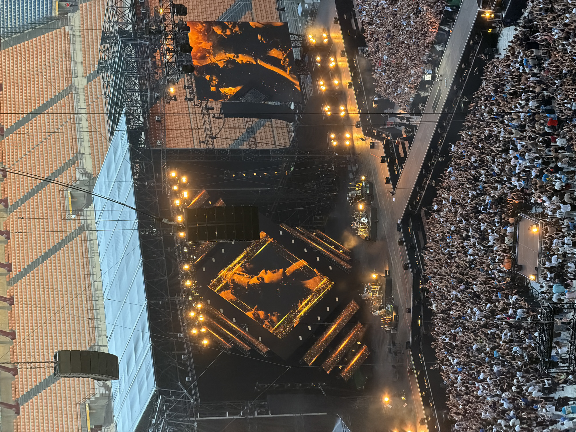 The image shows a large outdoor concert taking place in the San Siro stadium in Milan, Italy. A massive stage is set up with large screens displaying images of what appears to be a singer. Numerous spotlights illuminate the stage and the surrounding area. A huge crowd of people is gathered in front of the stage, many with their hands raised. The stands of the stadium are visible in the background, mostly empty but with some people seated. The overall scene suggests a lively and energetic musical performance.