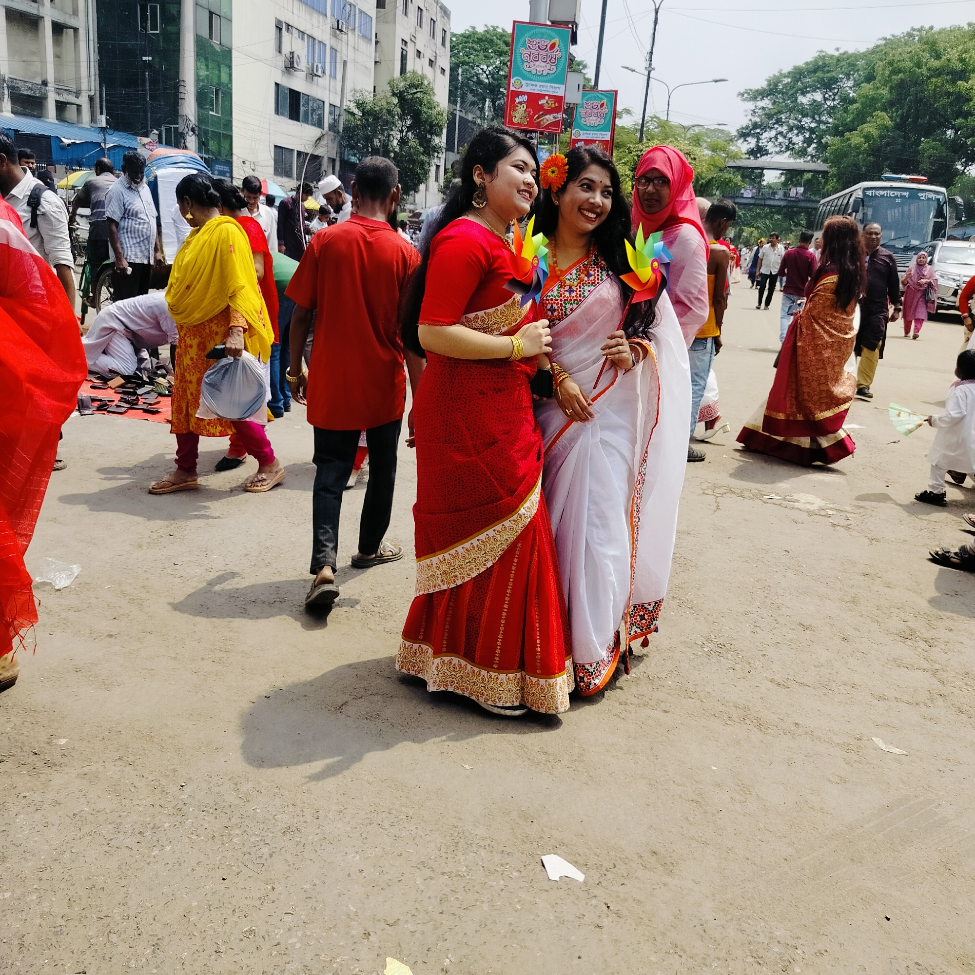 The image depicts a street scene in Dhaka, Bangladesh, likely during a cultural festival or celebration. Two young women in traditional sarees (one red, one white with red trim) are posing together and smiling, each holding a colorful pinwheel. They are surrounded by other people, many of whom are also wearing traditional attire. A man in a red shirt is walking away from the camera, slightly behind the women. In the background, there are buildings, street signs written in Bengali, and other pedestrians. A bus with "বাংলাদেশ পুলিস" (Bangladesh Police) written on its side is visible in the distance. The street surface is bare and dusty. There is litter on the ground.