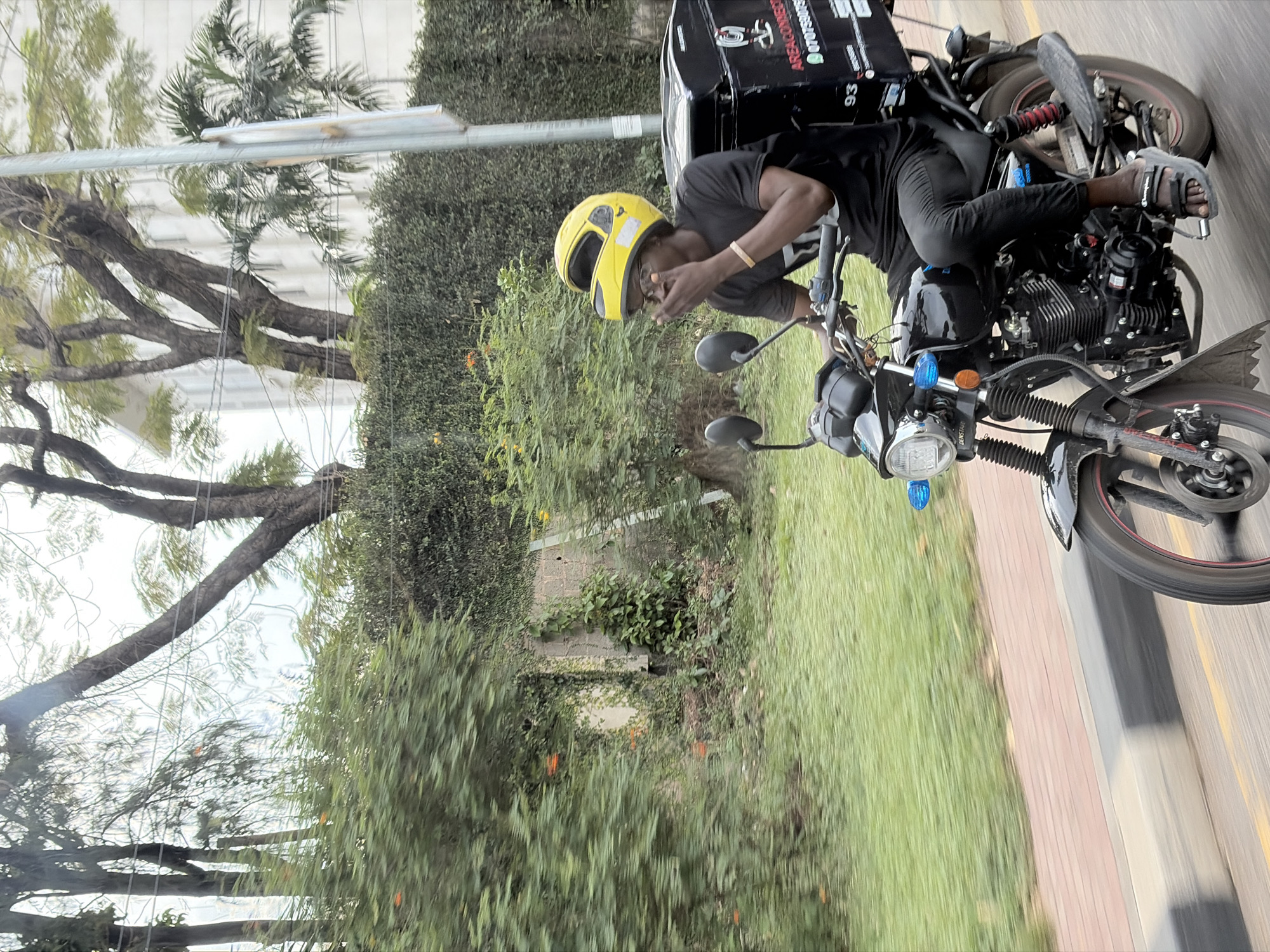 A man wearing a yellow helmet is riding a motorcycle with a delivery box on the back, presumably in Ikeja, Nigeria, given the context. The man is wearing a black t-shirt and pants, with sandals on his feet. He appears to be adjusting his helmet or glasses. The motorcycle is black with some red accents. The delivery box is black with white lettering. The background consists of trees, a building, a sidewalk, and a street. The image has a slight tilt, suggesting it was taken quickly or from a moving vehicle.