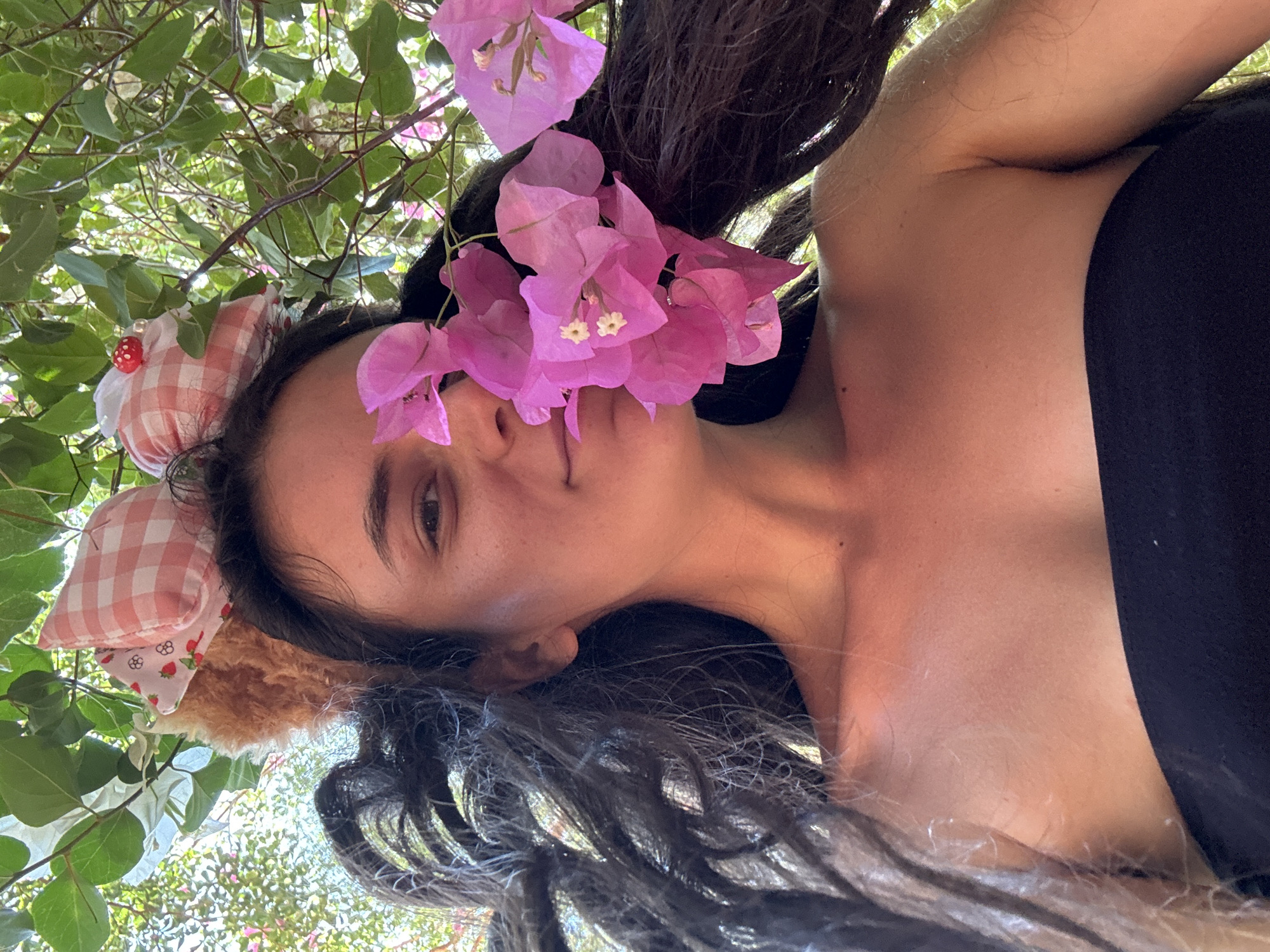 A young, light-skinned woman is pictured in an outdoor setting. She is lying on her back, looking up at the camera, partially framed by greenery and pink bougainvillea flowers. The background is bright and seems to be a garden or wooded area. She is wearing a black strapless top. She has dark hair, with some strands framing her face. She wears a pink gingham bow with white trim and a small mushroom embellishment on the side, suggesting a playful or whimsical theme. The picture is taken from a low angle, looking up towards her, which creates a sense of immersion in the natural surroundings. The location, Mũi Né, Vietnam, is not evident from the image itself.