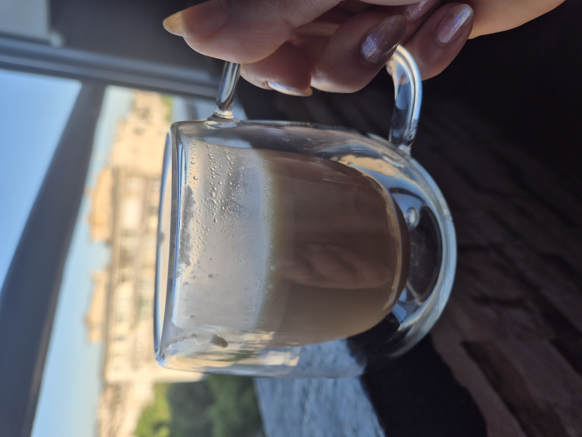 A close-up shot depicts a hand holding a clear glass mug containing a layered beverage, likely coffee with milk or creamer, in front of a window offering a cityscape view. The beverage appears to have a distinct separation between a frothy, lighter layer and a darker, coffee-colored base. The hand holding the mug has manicured fingernails. Outside the window, the buildings and greenery suggest a residential or commercial area within a city. The exact location in the city of Unknown, Unknown cannot be determined from the image.