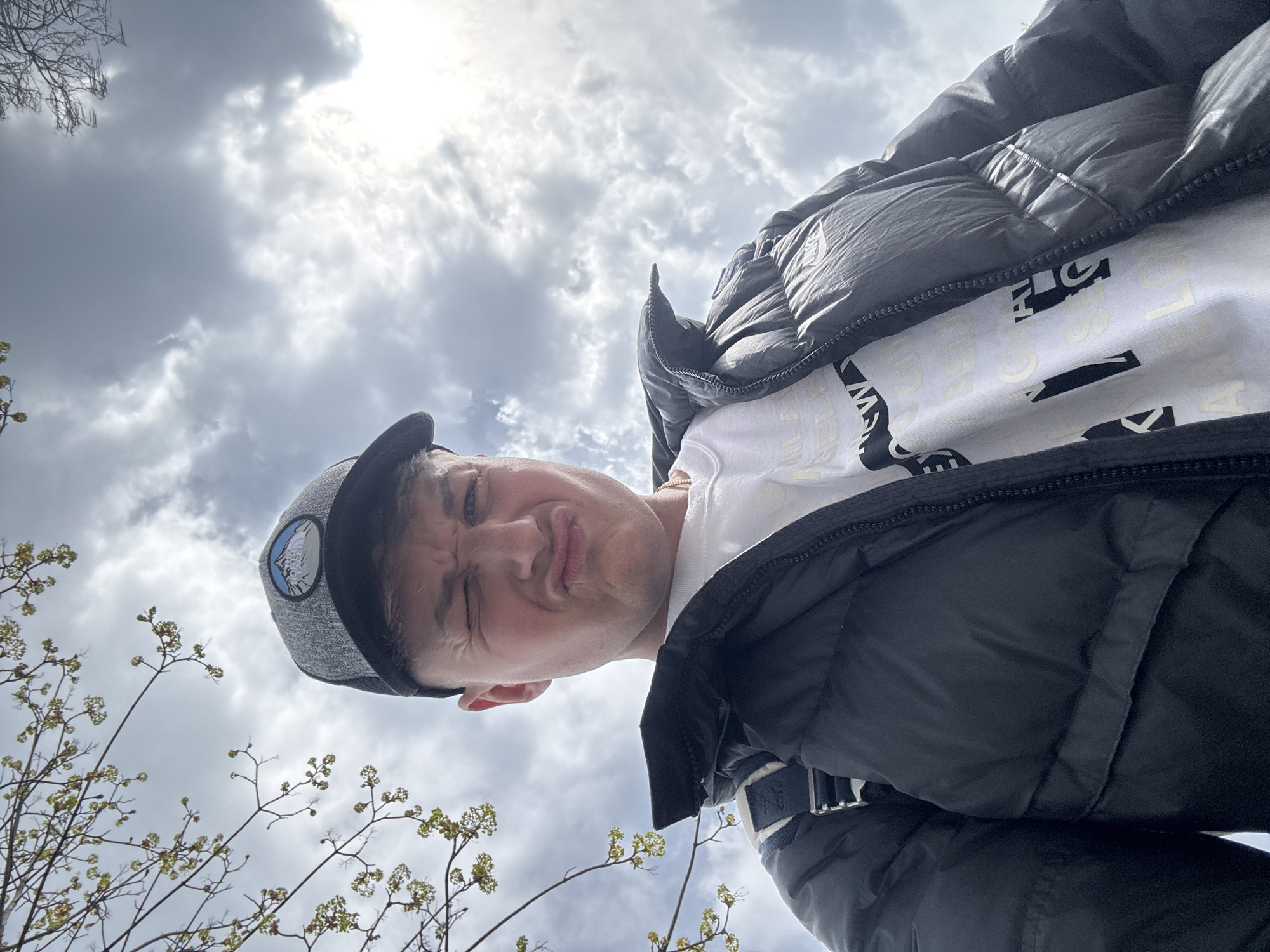 A low-angle outdoor selfie shows a person wearing a grey cap with a circular logo, a white t-shirt with black lettering, and a black puffer jacket. The person is making a funny face. A cloudy sky is visible in the background. In the lower-left corner, bare branches with small green buds are visible. The location is uncertain, but the picture may be taken in a park in Plovdiv, Bulgaria.