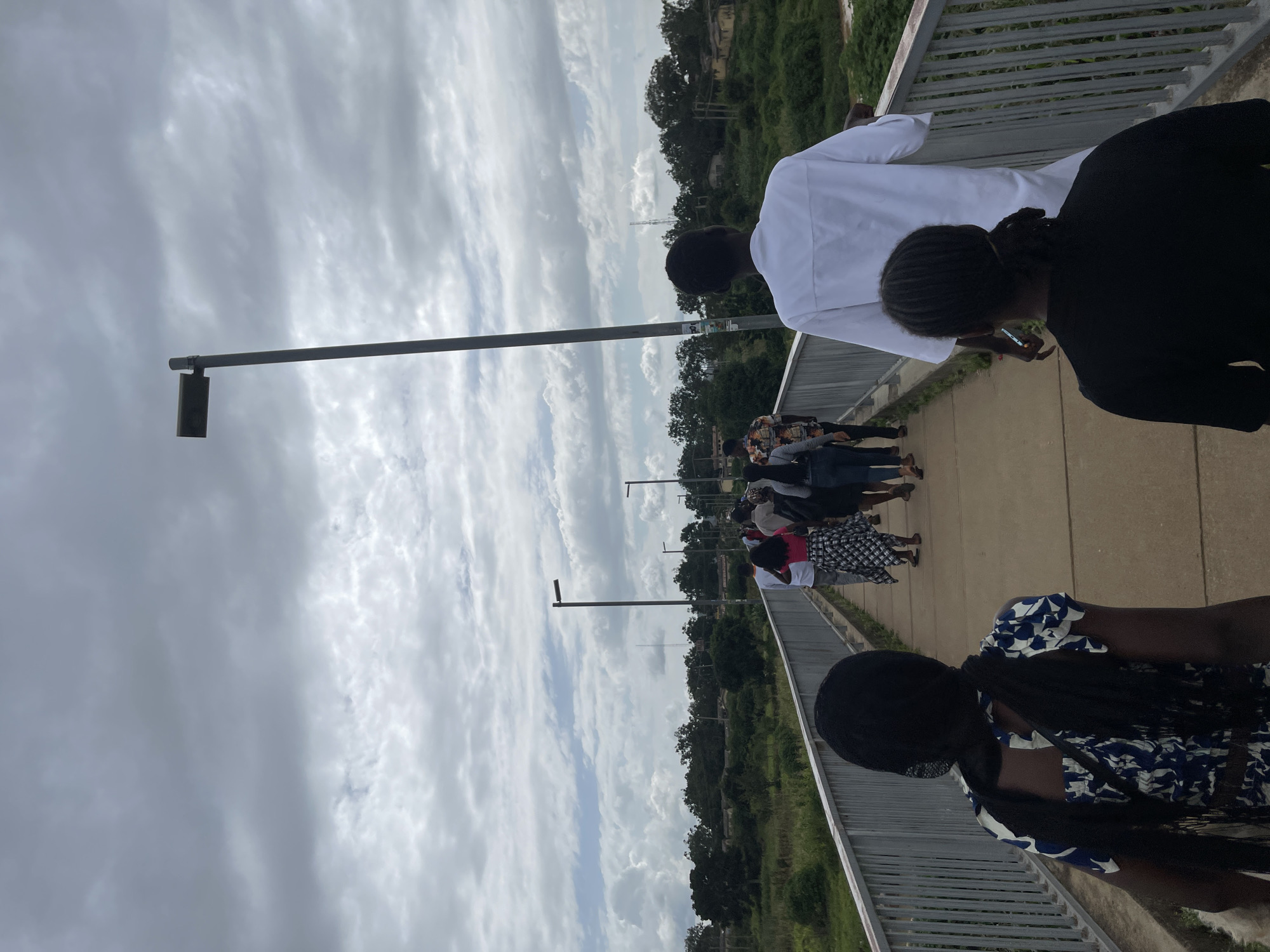 In Rafin Par, Nigeria, a group of people are walking across a pedestrian bridge under a cloudy sky. The bridge is made of concrete with a metal railing. The people are walking in the same direction, away from the camera. The person closest to the camera is a woman wearing a black headscarf and a blue and white patterned dress. Behind her, a man in a white robe walks forward, his back to the camera. Further ahead, other individuals are visible, their clothing varied. The surroundings include lush green vegetation and trees lining the landscape, with some buildings in the distance. Two lampposts are present on the bridge, one in the foreground and another further down. The overall scene suggests a casual daytime activity.
