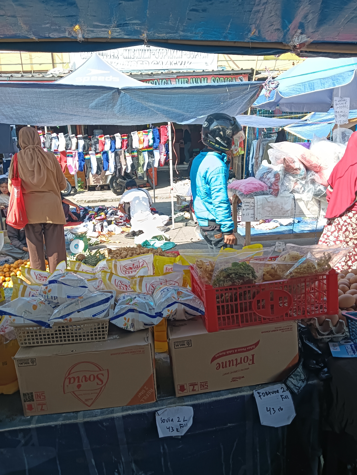 The image depicts an outdoor market scene in Katapang, Indonesia, likely a roadside market under tarps. In the foreground, there are several boxes and baskets containing food items. One large box on the left is labeled "Sovia" and indicates "Refill 2 liter". Another box next to it is labeled "Fortune" and contains cooking oil. A red plastic crate holds various packaged snacks. To the right of the crate, a large quantity of brown eggs are visible in cardboard trays.
In the middle ground, a woman wearing a hijab and a brown cardigan stands facing away from the camera, holding a red bag. To her left, a child is partially visible. A man in a white outfit is attending to goods spread on the ground. There is a scale visible near him. Behind them, a rack displays an assortment of colorful socks.
The background shows more market stalls with various goods, including clothing and other items, covered by blue and gray tarps. A motorcycle is parked behind the sock display. The overall impression is of a bustling, informal marketplace with vendors selling a variety of products. The lighting suggests it is daytime.