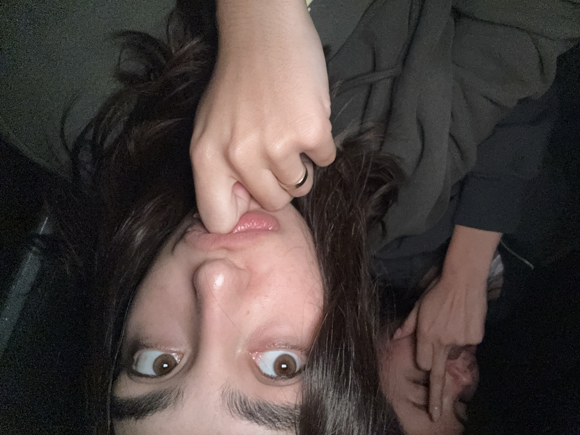 A selfie of a person with dark hair and brown eyes is oriented upside down, with a hand on the upper left side of the image pulling on the person's lip. A gold ring is visible on the hand. Another person’s face is partially visible on the lower right, with a hand near their face. The background is dark and indistinct. There is no indication of the city of Puerto Marqués, Mexico, in this image.
