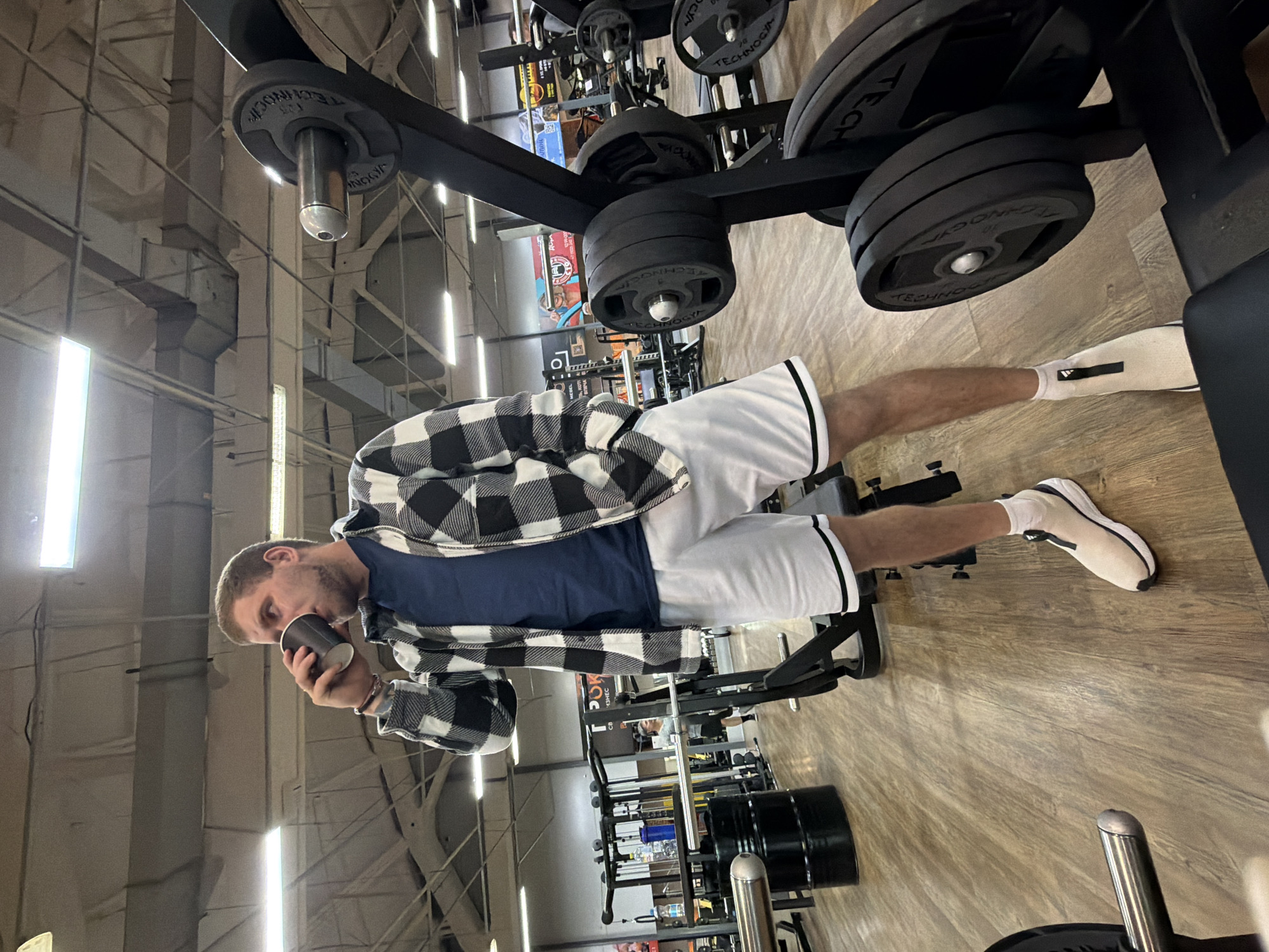 A man is standing in a gym, drinking from a cup. He is wearing white shorts with black trim, a navy blue t-shirt, a black and white plaid jacket, and white sneakers. The gym appears to be in the city of Chaikovsky, Russia, based on a storefront sign in the background. Various exercise machines and weights are visible around him.
