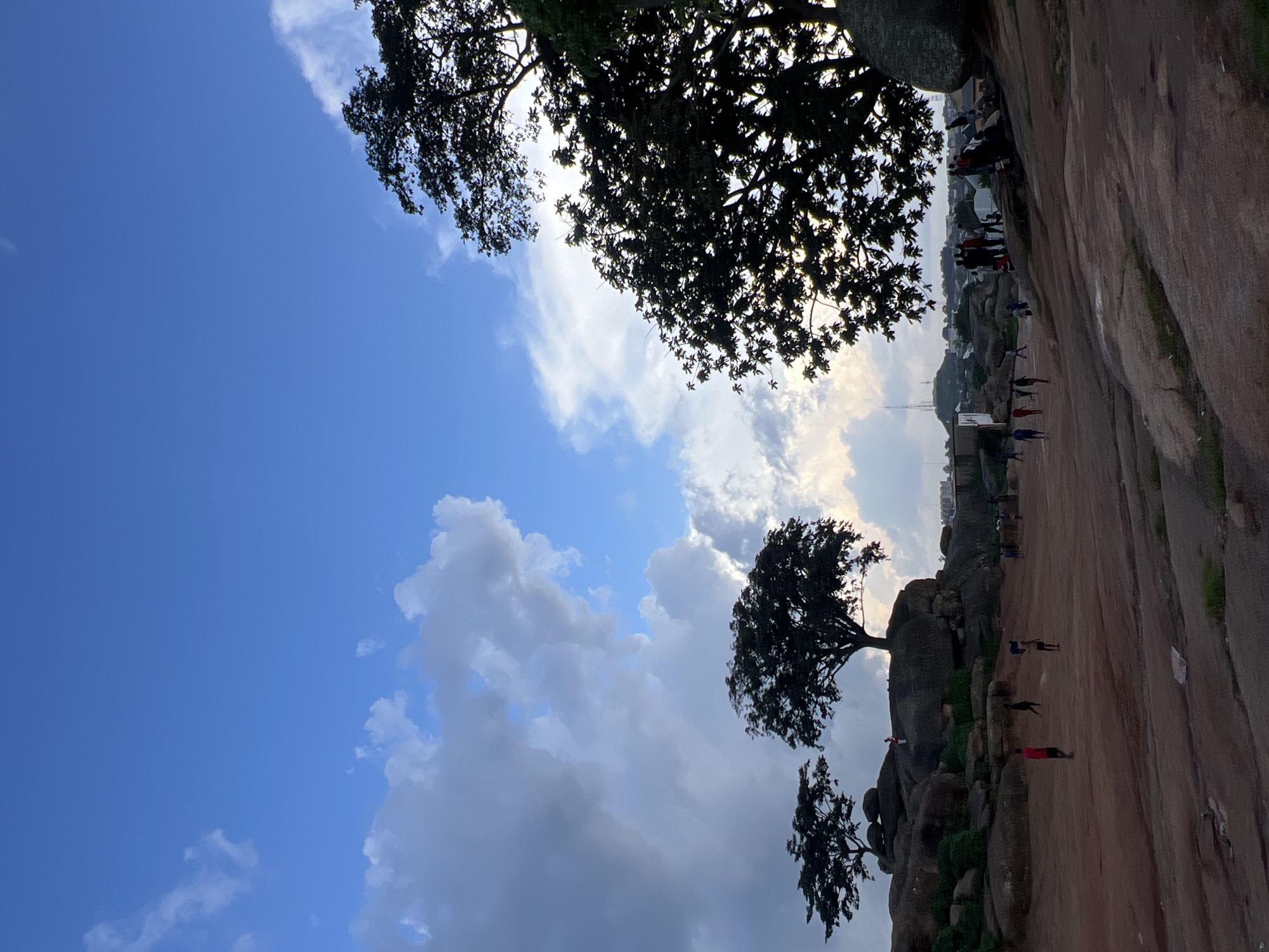 This media file, taken in Jos, Nigeria, depicts a group of people gathered on a rocky landscape under a partly cloudy sky. The scene appears to be a natural park or a recreational area known for its large rock formations. Several individuals are visible, some walking or standing on the rocky terrain, while others are seen in the distance near some buildings. The time of day appears to be late afternoon or early evening, given the warm, slightly diffused sunlight and the shadows cast by the trees and rocks. The weather seems pleasant, with a mix of blue sky and scattered clouds, suggesting a generally fair day.
The foreground is dominated by the texture of the large rock formations, with some sparse vegetation growing in the crevices. Silhouetted trees are prominent against the sky, adding a natural element to the scene. In the background, a town or city is visible, with buildings and infrastructure suggesting an urban or semi-urban environment bordering the natural landscape. The people present are casually dressed, and their activities suggest leisure or a gathering. Some individuals are positioned on a flat, sandy area that could be a designated pathway or gathering spot.
There is no visible text in the image. The overall impression is one of a relaxed, outdoor setting where people are enjoying the natural scenery and the company of others. The rocks and the open sky evoke a sense of space and natural beauty, while the distant buildings provide context for the location.