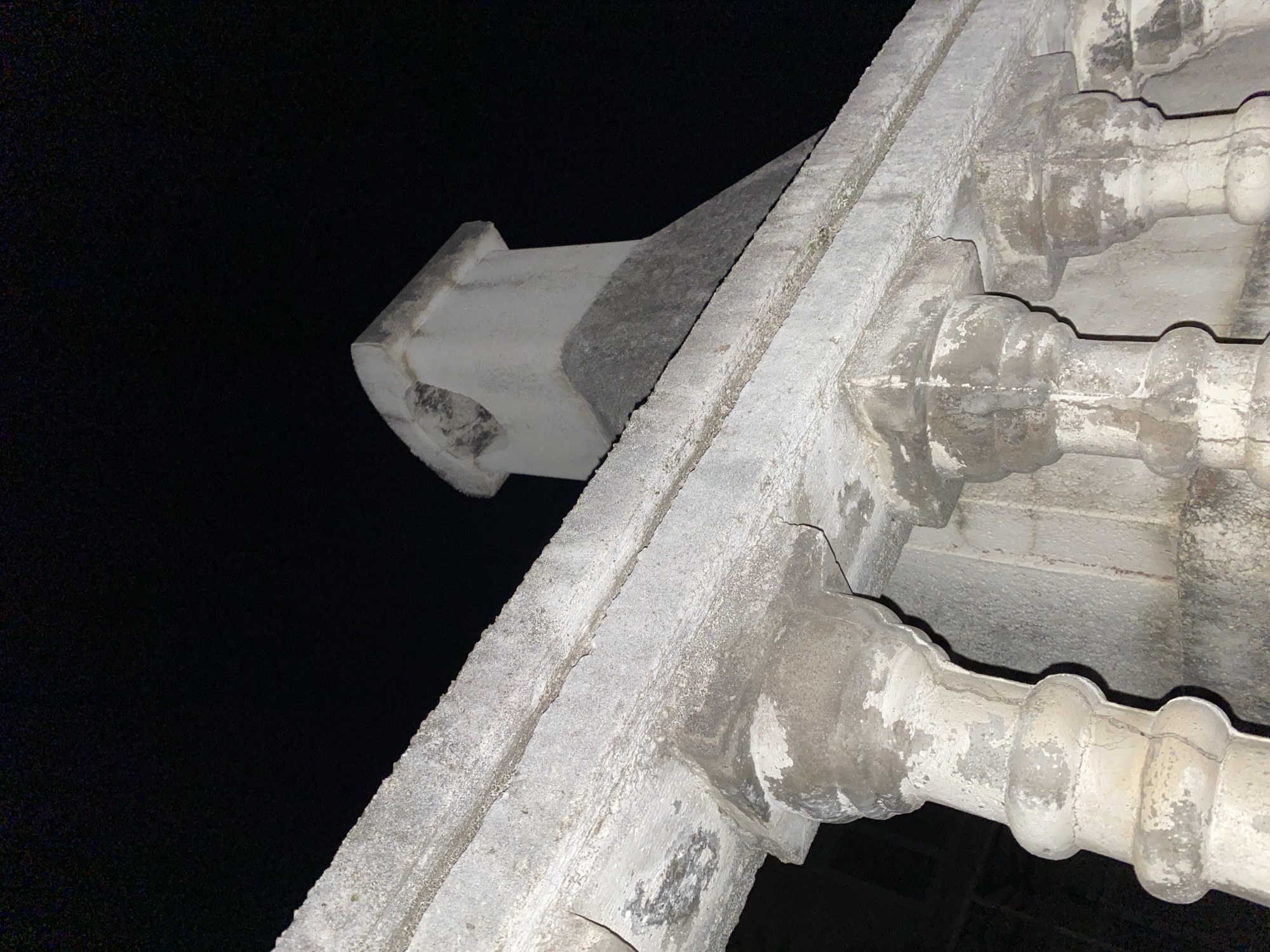 The image depicts a close-up, low-angle shot of a weathered, concrete structure against a dark, possibly nighttime sky. The structure features decorative elements, including molded bulbous forms separated by narrower sections, and a protruding, square-ended element at the top left. The surface of the concrete shows signs of age and wear, including discoloration and cracks. The location is likely within the city of Budva, Montenegro, based on the architectural style, but cannot be confirmed from the image alone. The image appears to focus on architectural details, and no people are visible.