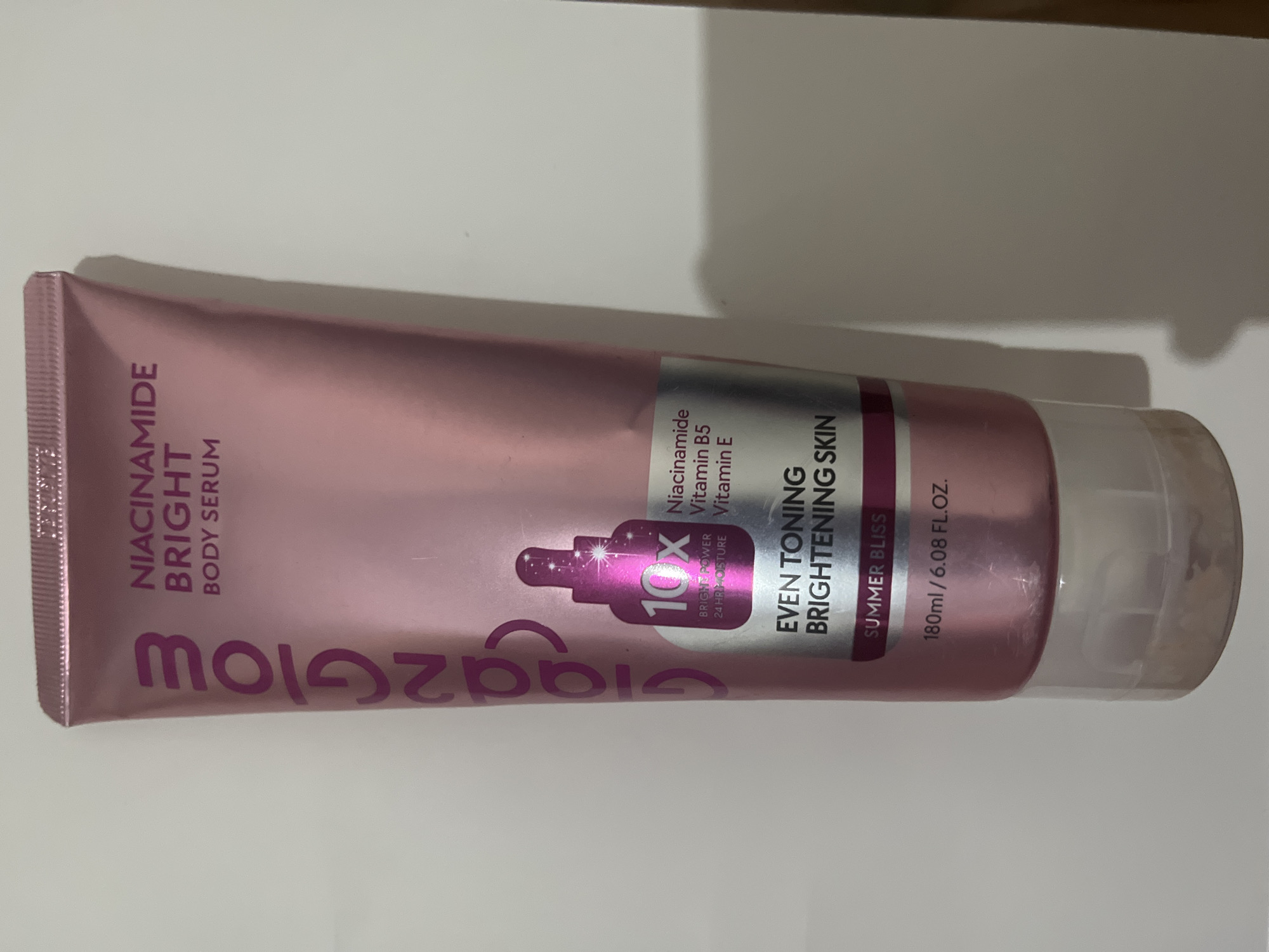 The image shows a pink metallic tube of "Glazeglow" Niacinamide Bright Body Serum. The tube is standing upright against a plain white background. The text on the tube indicates it contains "10x BRIGHT POWER 24 HR MOISTURE", Niacinamide, Vitamin B5, and Vitamin E. It is designed for "EVEN TONING BRIGHTENING SKIN" and has a scent called "SUMMER BLISS". The volume is listed as 180ml / 6.08 FL.OZ. The bottom of the tube is clear plastic, revealing some of the product inside. There are no people or other objects visible in the scene. The image does not contain any information about the location in Meliau Hulu, Indonesia.