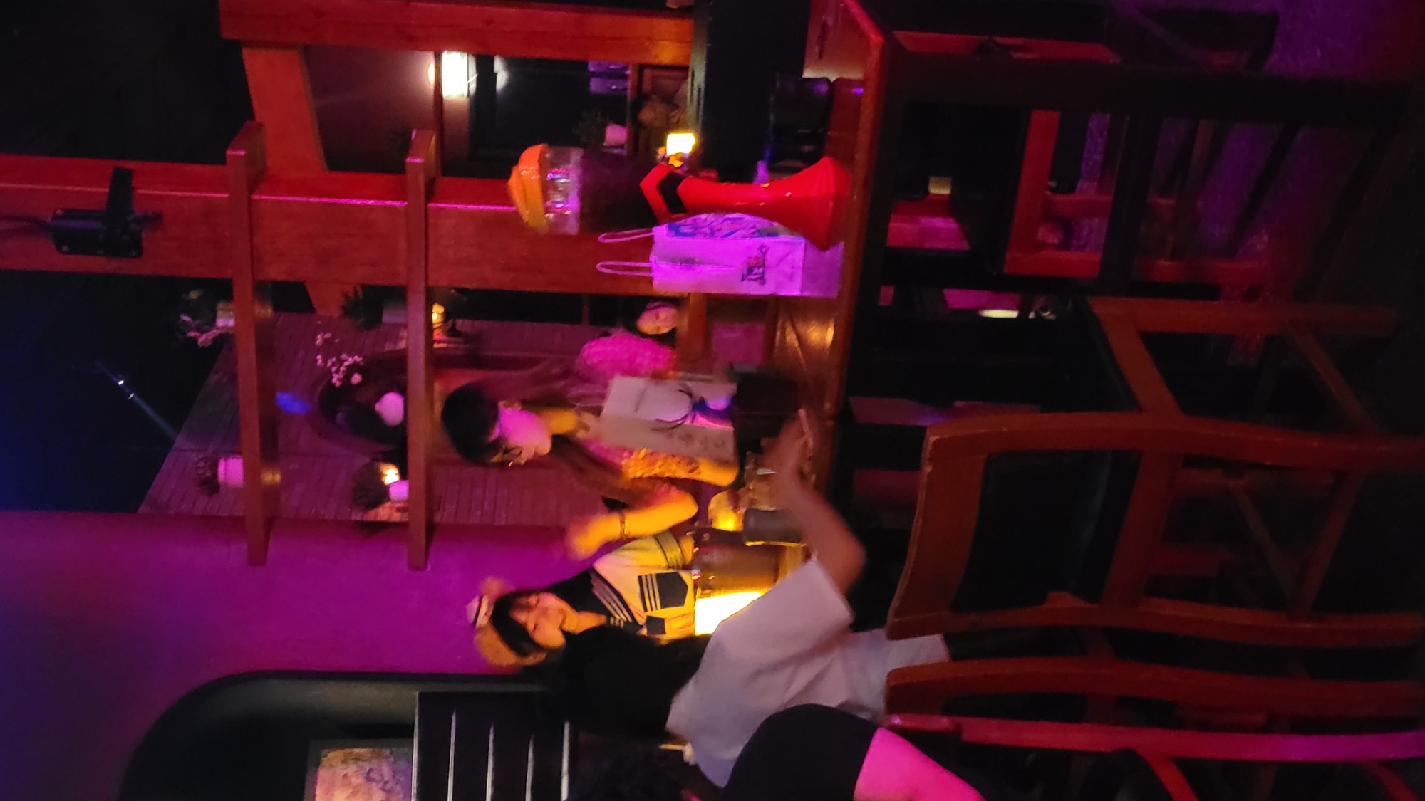 The image captures an indoor scene, likely a restaurant, bar, or cafe, at night or in the evening, bathed in striking purple and pink ambient lighting. Prominent wooden beams and structures, possibly part of the decor or furniture, frame the view.
Several individuals are present, appearing to be socializing or relaxing. In the foreground, a person with dark hair, wearing a white top and dark bottoms, is lying on their side on a surface, looking leftwards. Behind them, another person in a dark, possibly sailor-style top and a straw hat, is also oriented towards the left. Further back, a person wearing glasses is visible, holding what appears to be a menu or book. Another individual's head, topped with a hat, is partially seen in the background.
The setting features various objects, including a red megaphone, a yellow hard hat, and a white bag with faint, illegible text (possibly "I love...") placed on a wooden surface. Tables with bowls and small, glowing lights or candles are also visible, suggesting a dining or lounge area. The chairs have wooden frames. A black device, possibly a speaker or light fixture, is mounted on a beam. The overall atmosphere is dim and intimate, created by the colored lighting and wooden elements. The activity seems to be casual socializing in a relaxed environment.