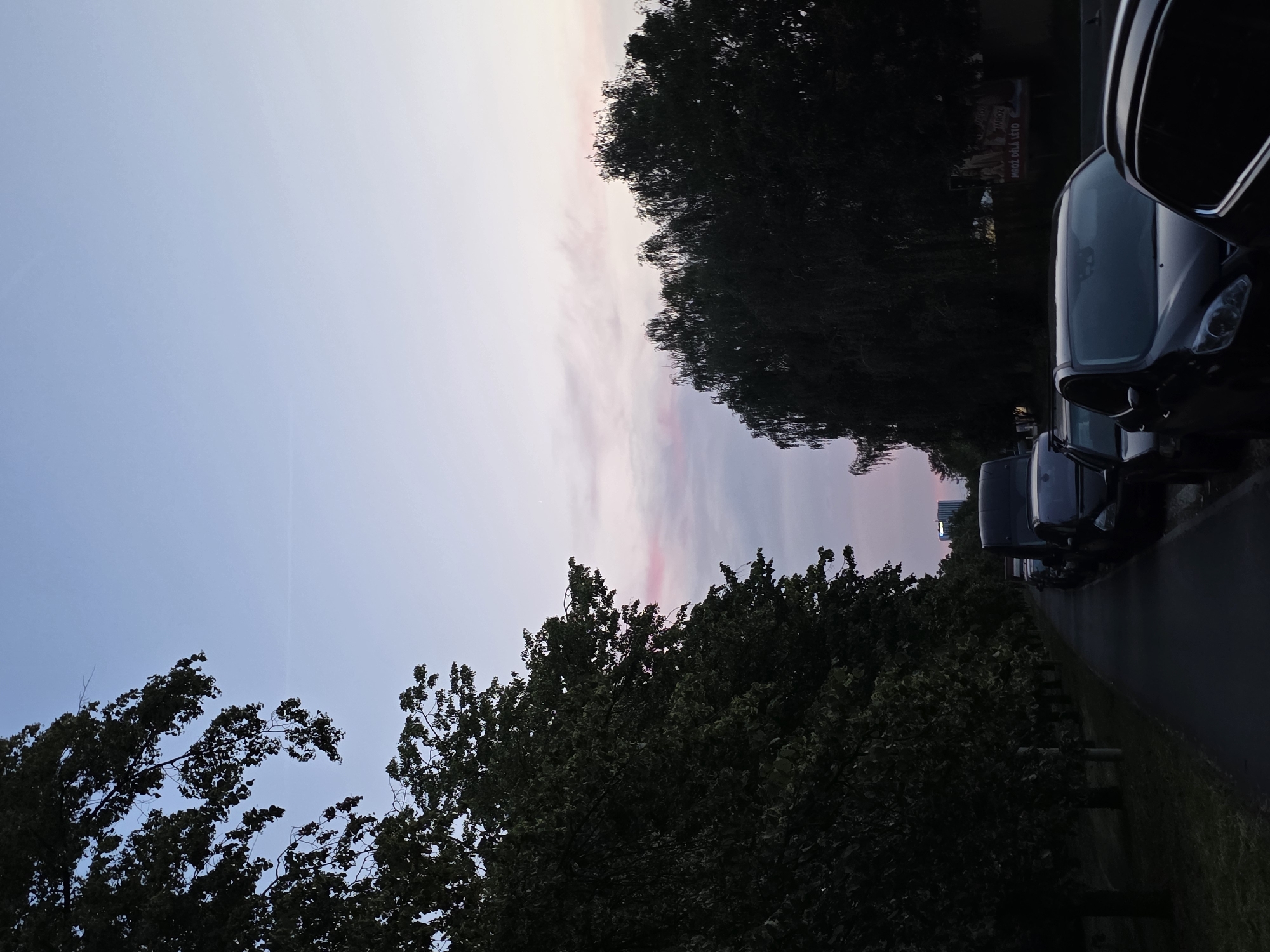 The image depicts a street scene at dusk in an unspecified urban location. On the left side of the frame, a paved pathway runs alongside a row of mature trees with dense foliage, casting long shadows. The pathway is bordered by a grassy area.
On the right side of the pathway, several cars are parked along the side of the road. The vehicles visible include a black van, a dark-colored SUV, and other cars whose details are less clear due to the low light conditions.
In the background, more trees are visible, forming a dark silhouette against the twilight sky. A distant building with a modern architectural design can be seen on the horizon, illuminated by the fading daylight. The sky exhibits a gradient of soft blues and pinks, indicative of sunset or sunrise, with a few faint clouds. A billboard with text is partially visible behind the parked cars. No people are present in the scene.