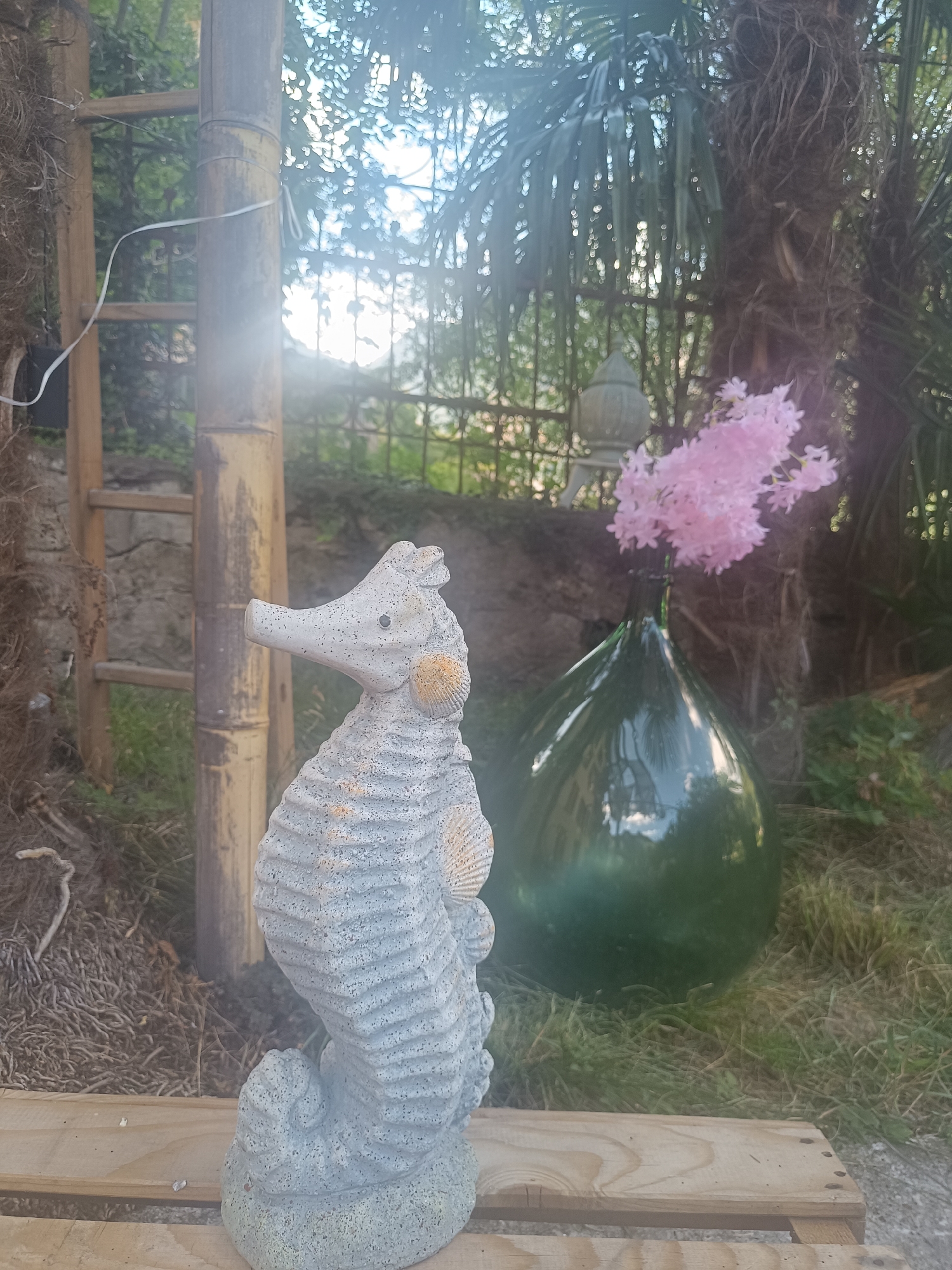 This is a medium shot, eye-level, outdoor photograph taken in Serravalle, Switzerland. The foreground features a light grey, concrete statue of a seahorse, positioned on a light-colored wooden pallet. The seahorse statue has textured detailing and a seashell accent on its side. To the left of the seahorse is a tall, bamboo pole structure with horizontal wooden rungs, resembling a ladder. Behind the seahorse and to the right, a large, dark green glass bottle is visible, containing a bouquet of small, pink flowers. In the background, there is dense foliage, including palm fronds, and a blurred, decorative metal fence. The lighting suggests it is daytime, with some lens flare from the sun. The environment appears to be a garden or an outdoor decorative space.