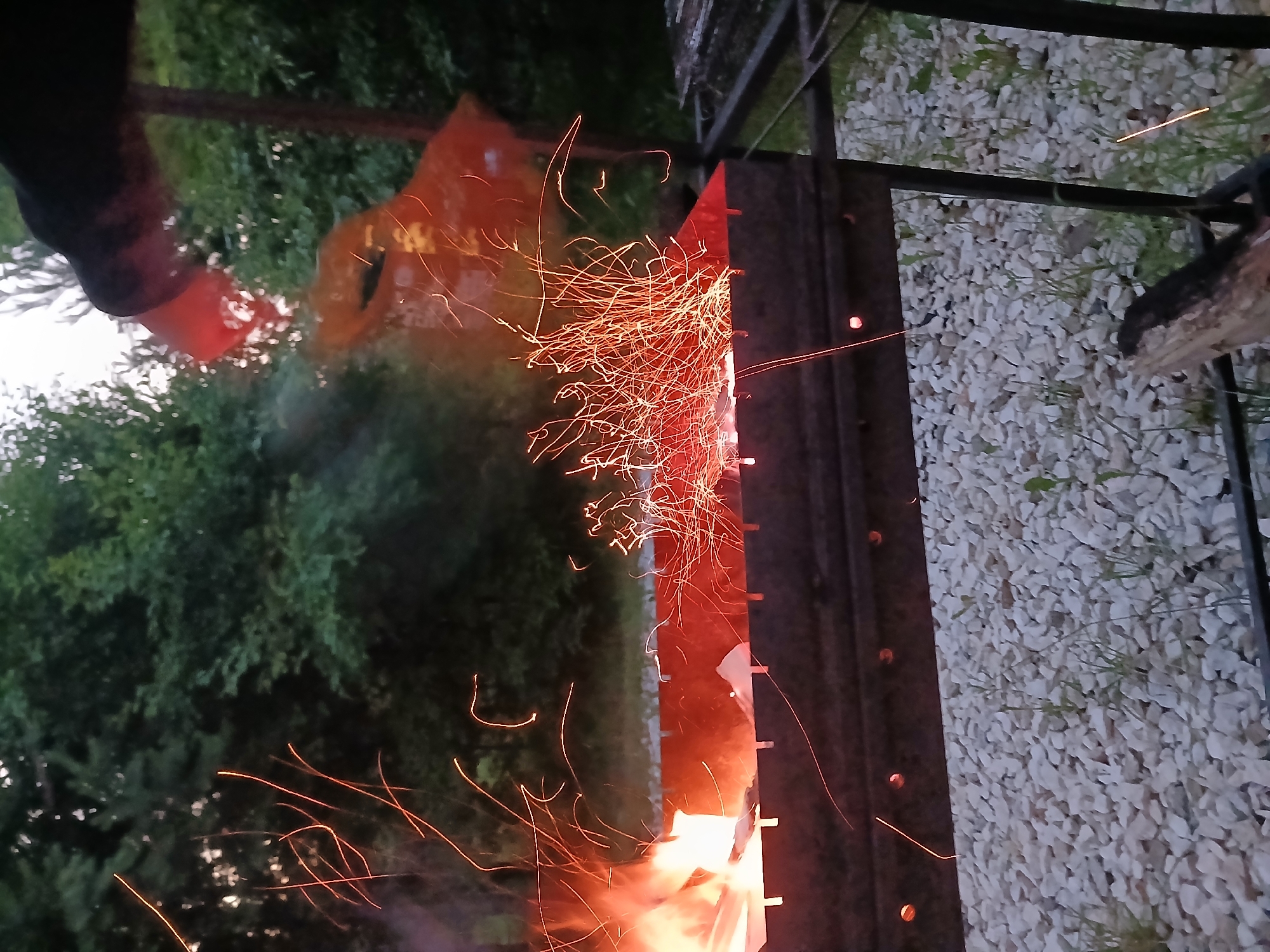 This media file shows a close-up, slightly blurry view of a fire being lit in an outdoor barbecue grill. The foreground is dominated by a dark metal barbecue grill with glowing embers and bright orange sparks flying upwards and outwards. The sparks create streaking patterns against a dark background, suggesting a long exposure or movement.
To the left, a person's arm and hand, partially obscured by shadow and the red glow of the fire, are visible. They appear to be holding or interacting with something that is contributing to the fire, possibly a starter or fuel. Behind the grill, lush green foliage from trees can be seen, indicating an outdoor setting surrounded by nature. The ground around the grill is covered in light-colored gravel and some sparse green grass.
The lighting and the presence of sparks suggest that it is either dusk or evening, as the fire is the primary light source in the immediate vicinity. The overall mood is one of warmth and activity, centered around the preparation of an outdoor fire for cooking or gathering. The image seems to capture a moment of lively ignition and the beginning of a fire. There is no visible text in the media file. The context provided places the scene in Verkhneye Dubrovo, Russia.
