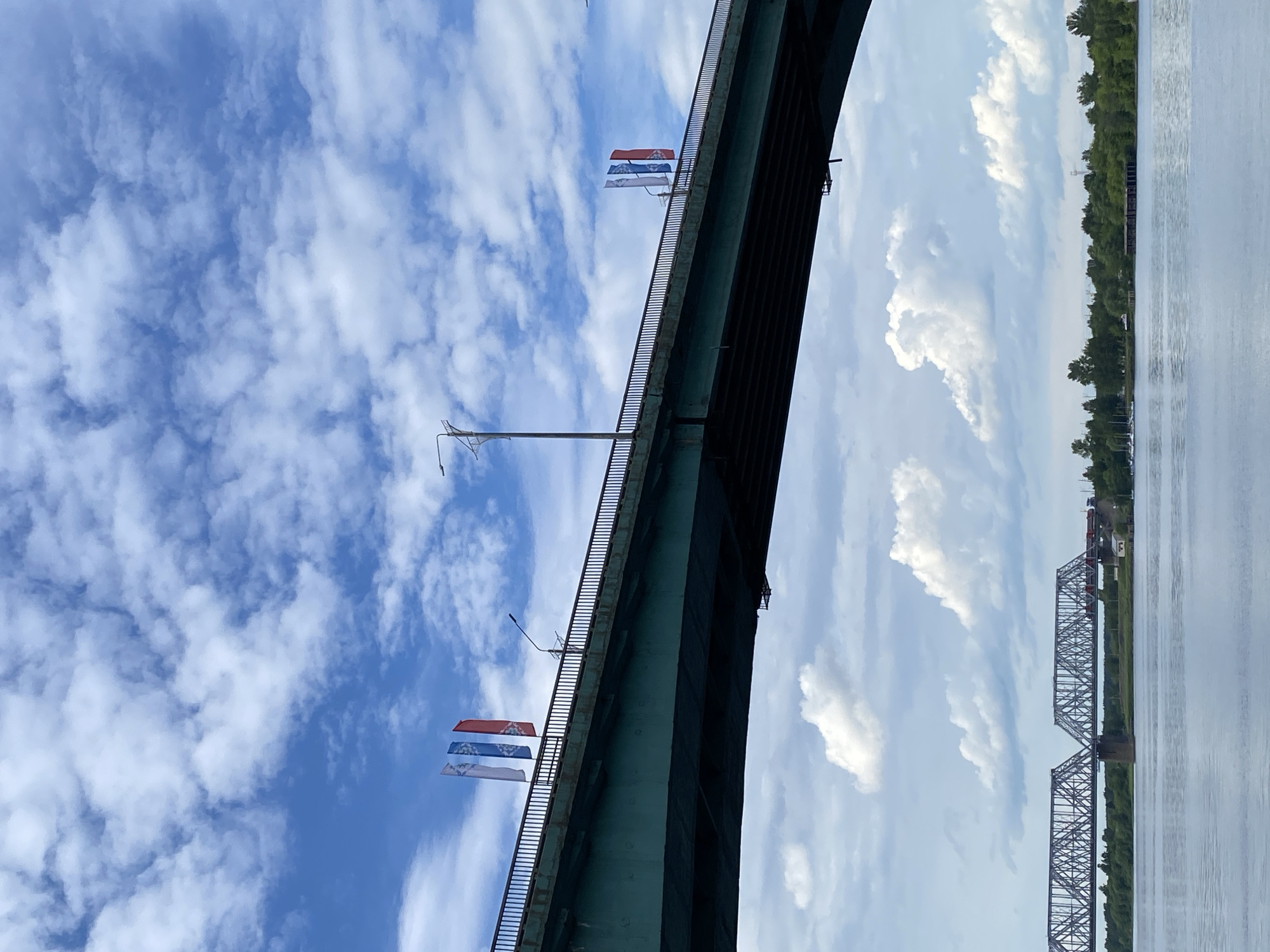 The image, originally rotated, presents a daytime view looking up towards a large bridge spanning a wide river in Заволжье, Russia. A substantial, dark green or gray vehicular bridge curves across the upper left and central parts of the frame. Two identical decorative banners hang vertically from the bridge's railing. Each banner features a red top section, a white middle section with an indistinct blue emblem, and a blue bottom section.
Below the main bridge, the wide, calm surface of a river, likely the Volga, stretches across the middle distance, reflecting the bright sky. On the far bank of the river, a dense line of green trees marks the shoreline. Further in the distance, a second, older steel truss bridge, likely a railway bridge, spans the river. The sky above is a vibrant blue, filled with scattered, fluffy white cumulus clouds, indicating fair weather. No specific activity or event is taking place. There is no discernible text visible, only the abstract blue emblems on the banners.