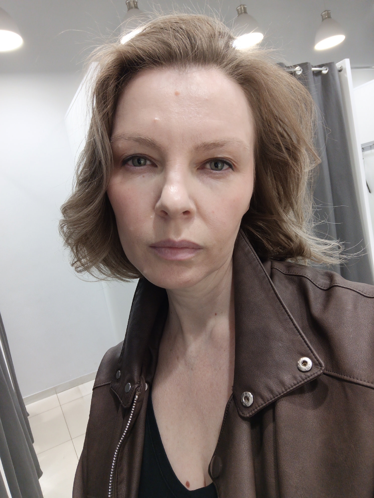 A woman, appearing to be in her 30s or 40s with light brown, wavy, shoulder-length hair and light green/hazel eyes, takes a selfie indoors in Chaikovsky, Russia. She wears a dark brown, possibly leather or faux-leather, jacket with silver snap buttons visible on the lapel, over a black top. Her facial expression is neutral and direct, as she looks straight into the camera. Minor blemishes are visible on her forehead and cheek.
The background suggests a fitting room or retail store setting, with plain white walls and grey curtains or drapes hanging from silver rods on both sides. Several bright artificial lights, including recessed spotlights and pendant fixtures, are visible on the white ceiling, illuminating the scene. A light-colored tiled floor is partially visible in the lower left corner. The image captures a personal moment, possibly while trying on clothing, within a commercial indoor environment. No visible text is present.