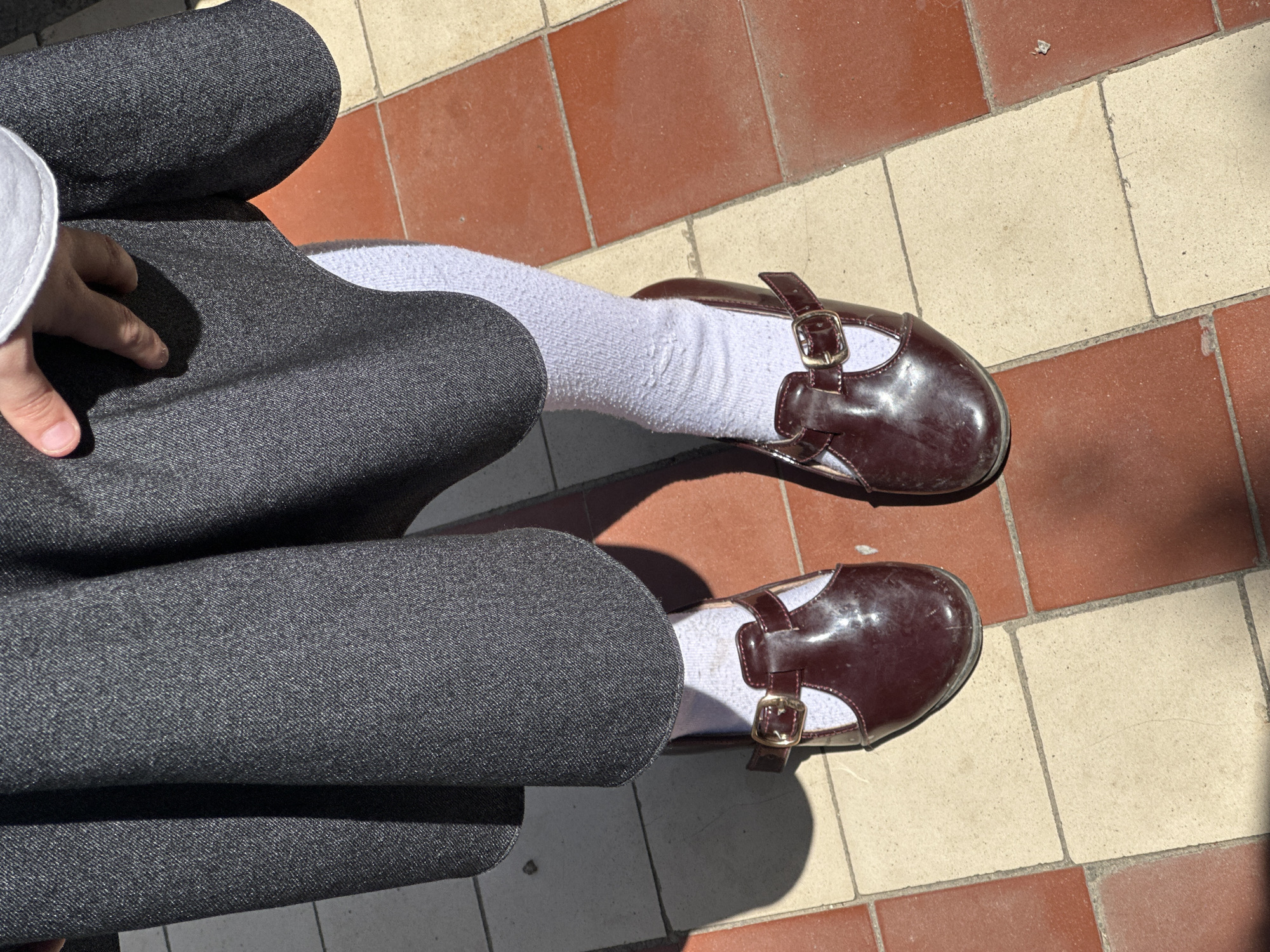 An overhead, eye-level shot captures the lower half of a person sitting or resting on a tiled surface in Agarak, Armenia. The subject is wearing dark gray trousers or a skirt made of a finely textured fabric, possibly part of a uniform, paired with white socks that are slightly wrinkled. Their feet are adorned with shiny, dark red or maroon Mary Jane-style shoes, featuring gold-colored buckles across the instep. The shoes appear scuffed and well-worn.
One hand, with neatly trimmed fingernails, rests gently on the person's lap, covered by the gray garment. The scene is brightly lit by direct sunlight, casting distinct, sharp shadows from the shoes and legs onto the ground, indicating it is daytime, likely mid-morning to afternoon.
The ground beneath the person is a checkerboard pattern of alternating reddish-brown and off-white square tiles. The tiles show signs of wear and dirt, with visible grout lines. No text is visible in the image.