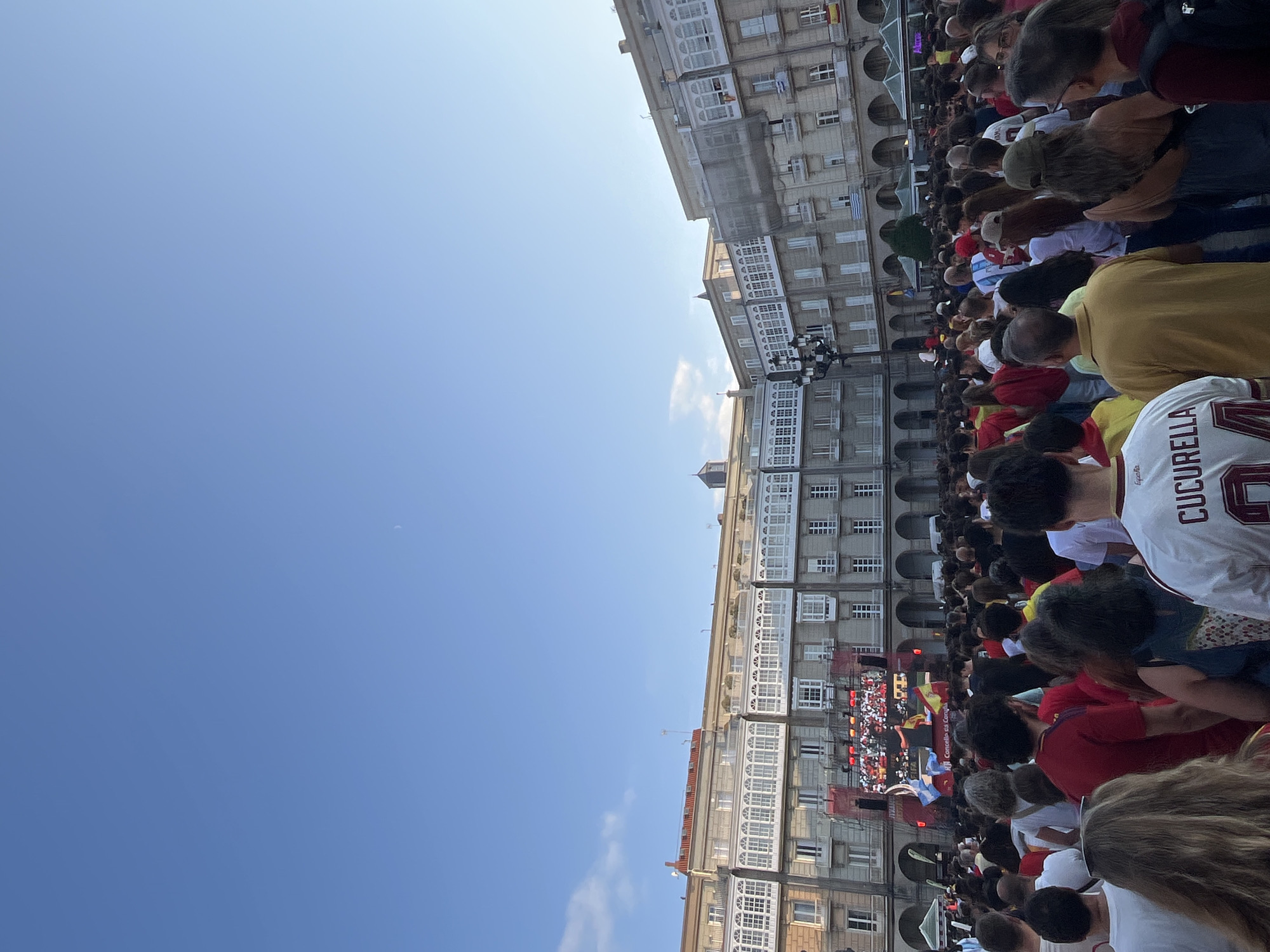The image shows a large crowd of people gathered in a public square in front of a large building with distinctive architectural features. The building has multiple floors with many windows and balconies, suggesting it is a historic or significant structure. In the center of the square, there is a large screen showing a broadcast, possibly a sporting event, with flags displayed in front of it. The people in the crowd are facing the screen, and many are wearing casual clothing, with some appearing to be wearing team colors or jerseys. The atmosphere seems to be festive, with a sense of shared experience among the attendees. The sky is clear blue, indicating a daytime scene. The architecture and style of the building, particularly the balconies, are characteristic of the city of A Coruña, Spain. This scene is likely taking place in the Plaza de María Pita, a central and prominent square in A Coruña, known for hosting public events and gatherings.