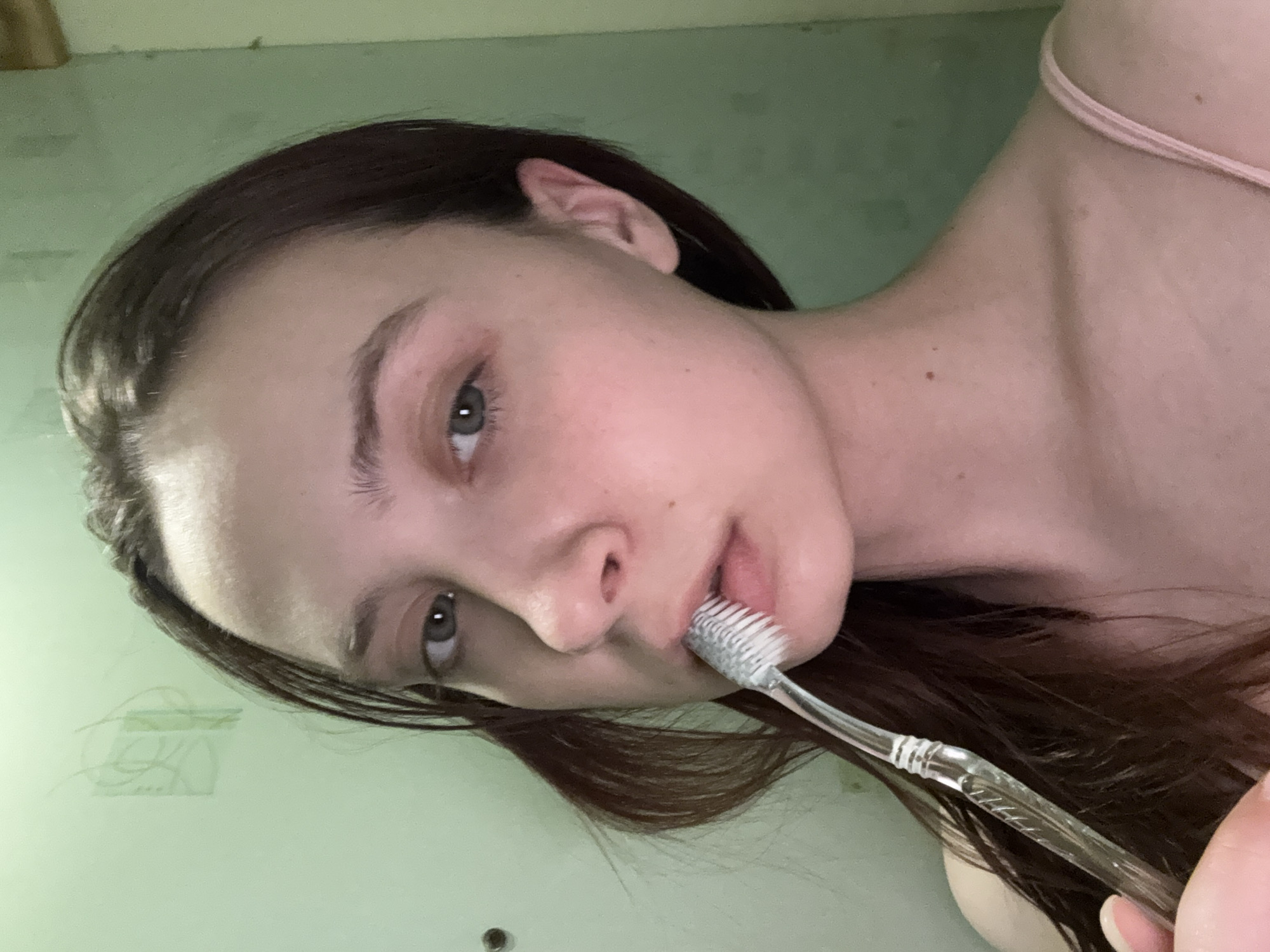 A young woman with long, dark hair and fair skin is holding a transparent toothbrush in her mouth. Her eyes are gray, and she is wearing a light pink tank top. The background is a light green wall. The location cannot be determined from the image.