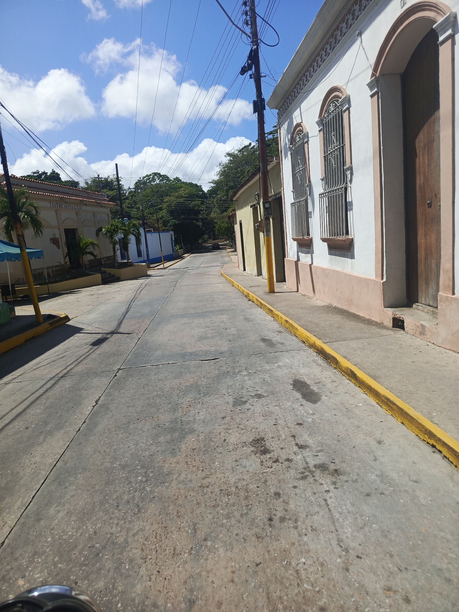 The image depicts a street in La Asuncion, Venezuela, showcasing a daytime scene with a clear blue sky dotted with scattered clouds. The street is paved and appears to be a low-traffic area, with visible cracks and wear on the surface. The right side of the frame features a building with a white facade and light pink accents, characterized by arched windows with decorative iron bars and a large wooden door. A yellow curb runs along the edge of the sidewalk adjacent to the building. Utility poles with numerous power lines are visible along the street, adding to the urban setting. On the left side of the street, another building labeled "Teatro Santa Lucia" can be seen, with trees lining the street further ahead. The overall scene conveys a quiet, sunny day in a historical urban area.