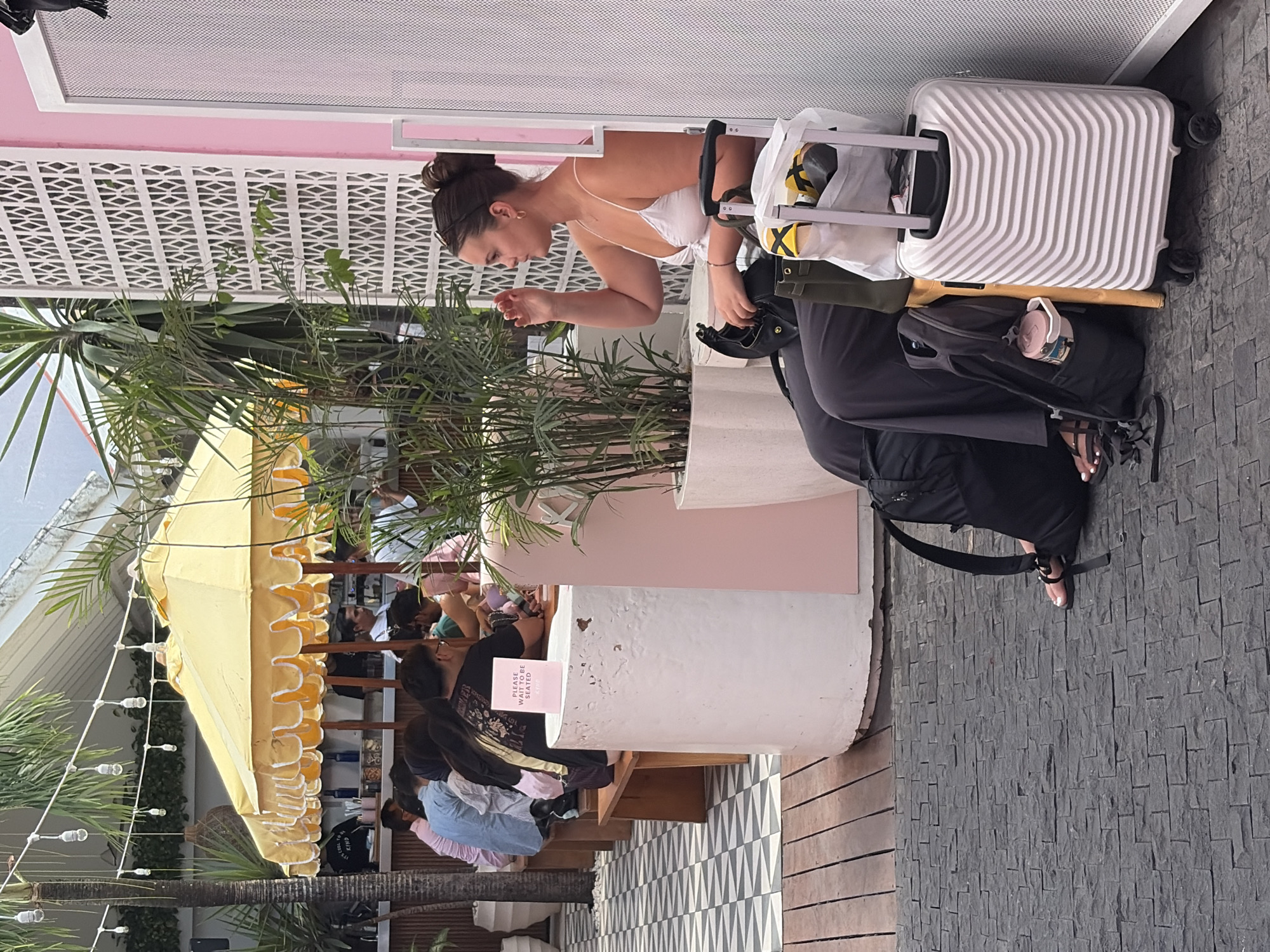 In North Kuta, Indonesia, a young woman with her hair pulled back in a bun, wearing a white tank top and dark pants, sits on a wooden bench. She is looking to her right and appears to be gesturing with her right hand. Beside her, on the ground, are two suitcases, one white rolling suitcase with a striped pattern and a black backpack. In front of the bench, on a circular white pedestal, is a pink sign that reads "PLEASE WAIT TO BE SEATED". Behind the woman, there are other people seated at tables, some under a yellow umbrella. The environment appears to be an outdoor seating area with plants and patterned flooring.